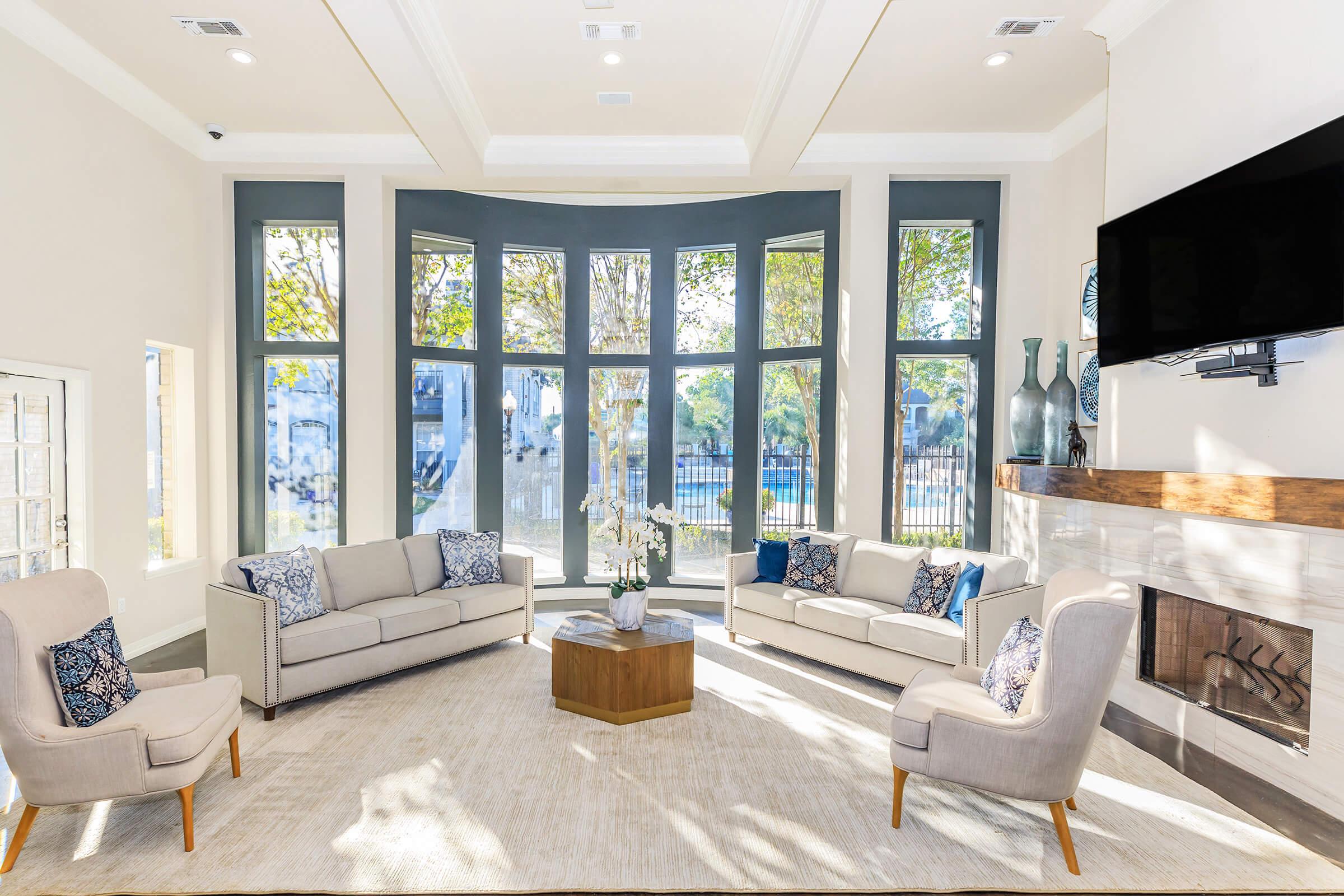 A bright and modern living room featuring a curved wall of large windows, light-colored furniture including two sofas and two armchairs with decorative cushions, a central wooden coffee table, a light area rug, and a wall-mounted television above a stylish fireplace. Natural light fills the space.
