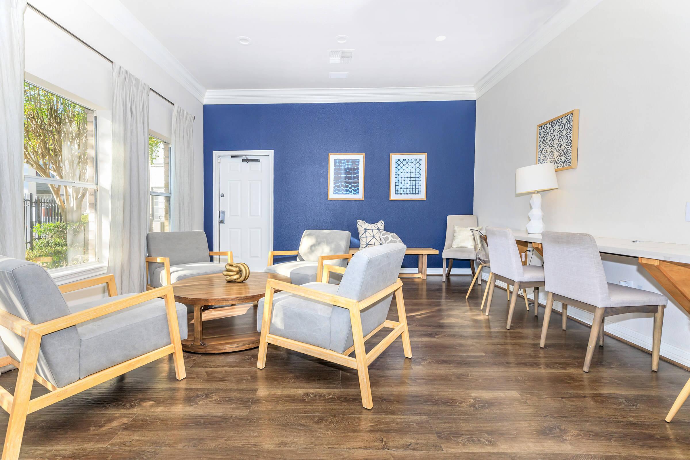 Cozy indoor seating area featuring light wood furniture with gray cushions, a round wooden coffee table, and a decorative lamp. The walls are a mix of blue and white, with large windows allowing natural light. There are decorative wall art pieces and a stylish, modern atmosphere.