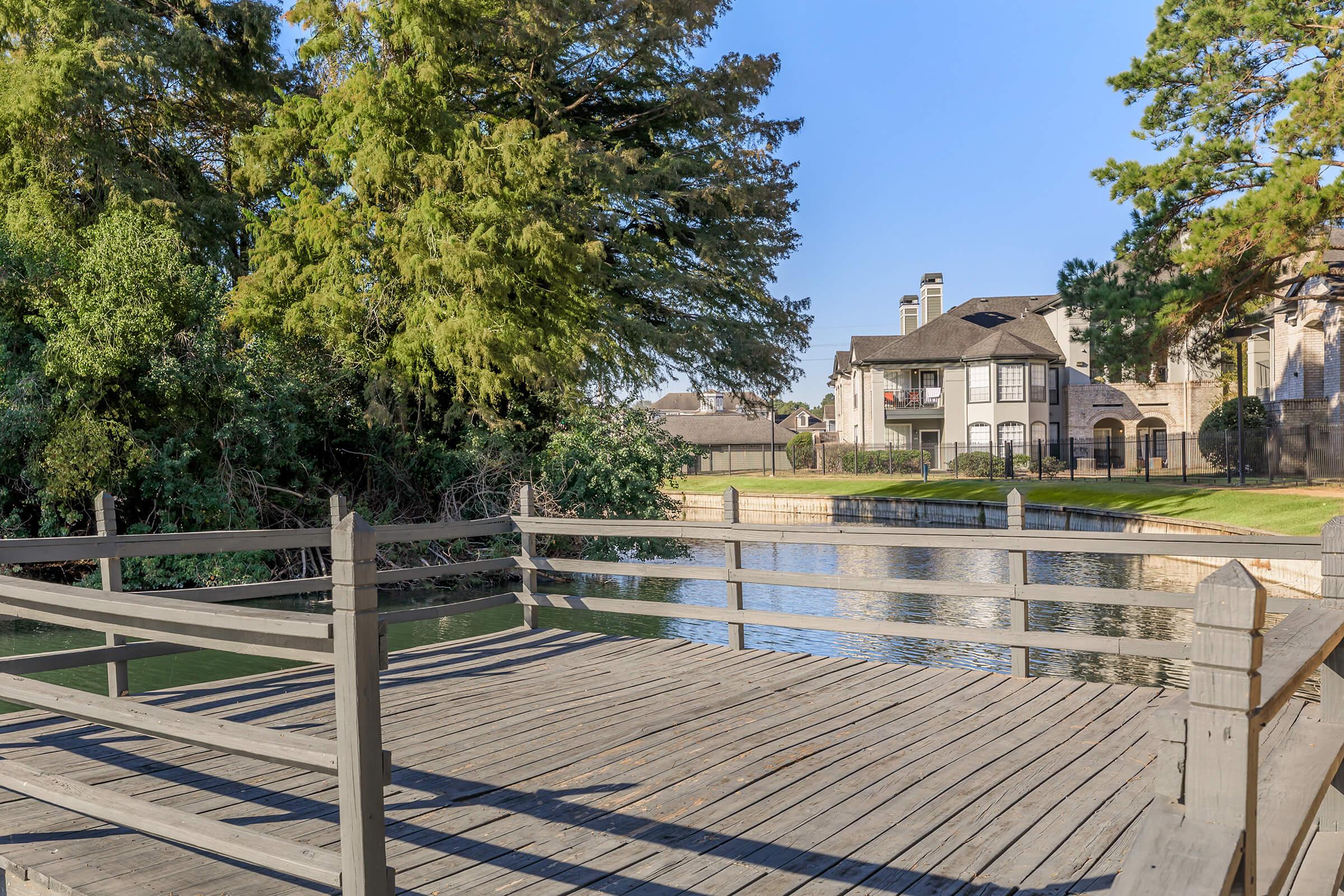A wooden dock extending over a calm body of water, surrounded by lush greenery. In the background, there's a modern residential building. The scene is illuminated by clear blue skies, indicating a sunny day.