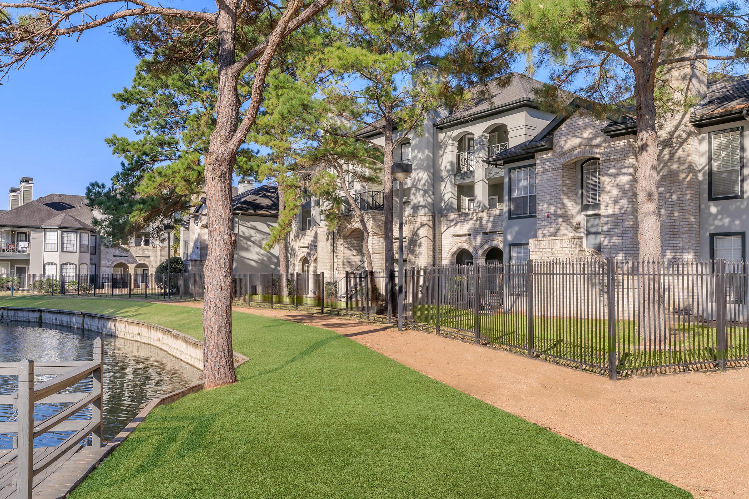 A serene landscape featuring a lakeside path lined with trees and well-maintained lawns. In the background, landscaped apartment buildings with a mix of brick and modern architecture. The scene is bathed in sunlight, creating a peaceful atmosphere.