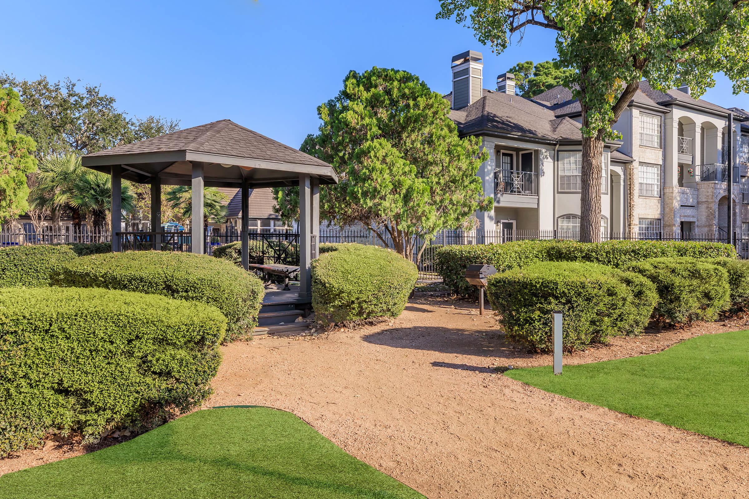 A serene outdoor space featuring a gazebo surrounded by well-maintained shrubs and grassy areas. In the background, an apartment building is visible, framed by trees under a clear blue sky. The scene conveys a peaceful environment suitable for relaxation.