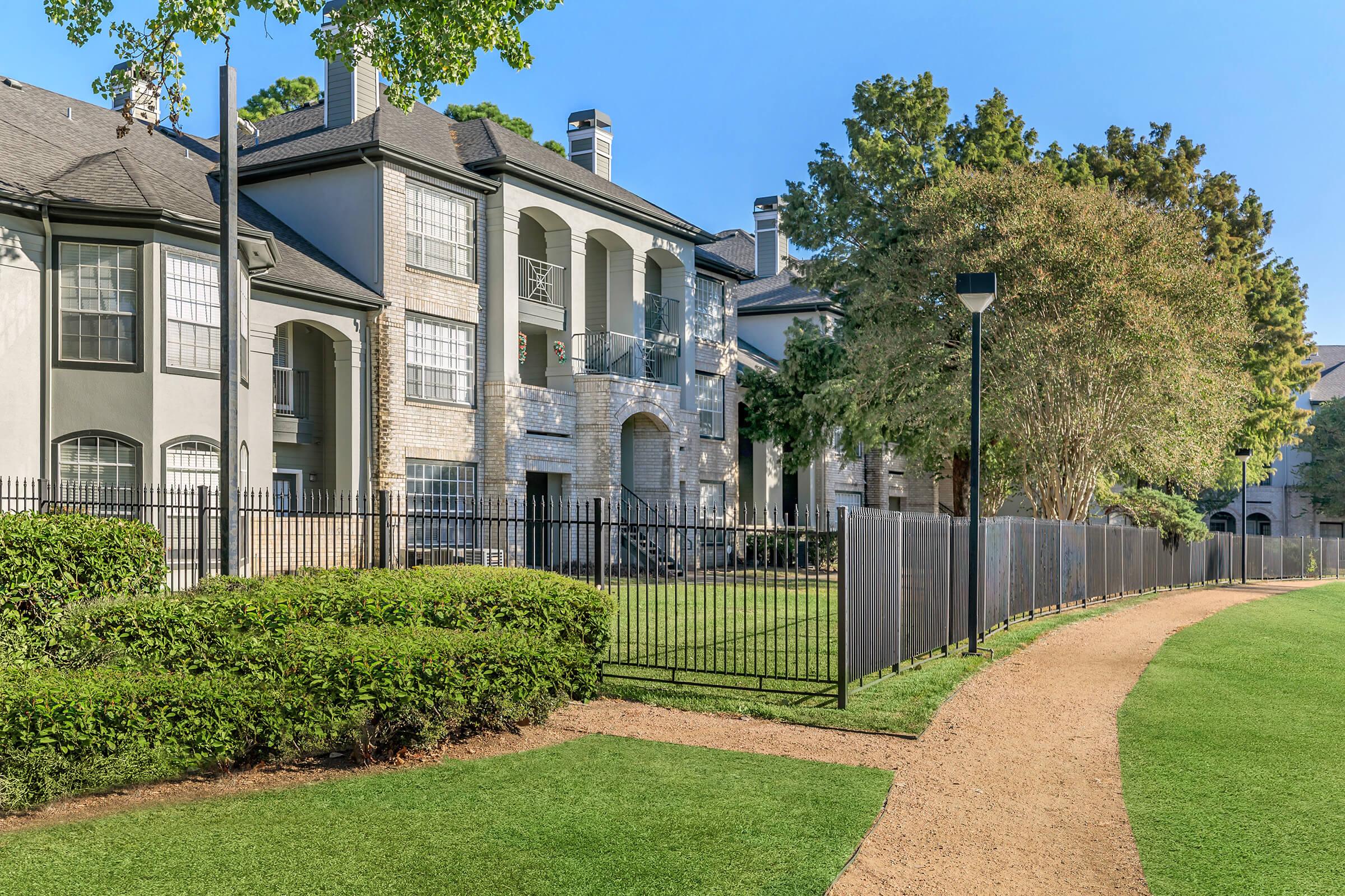 A well-maintained residential area featuring two-story apartment buildings with a mix of brick and stucco exteriors. Lush green grass and neatly trimmed bushes line a walking path that runs alongside a black metal fence, with trees providing shade under a clear blue sky.