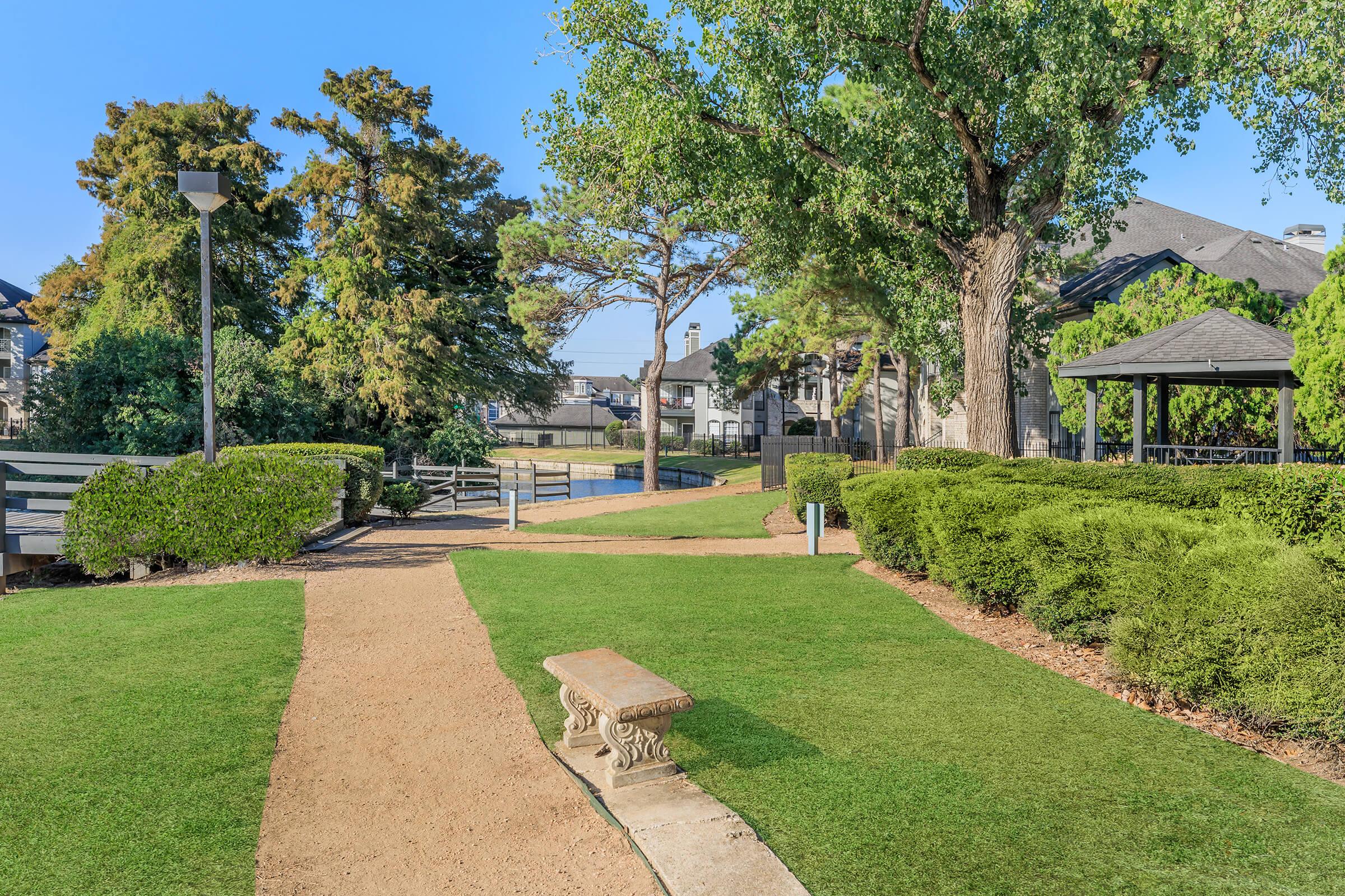 A serene park pathway with lush green lawns, neatly trimmed bushes, and a bench. In the background, a gazebo and trees provide shade, while a small water feature can be seen. Bright blue sky enhances the peaceful atmosphere, making it an inviting outdoor space for relaxation and leisure.