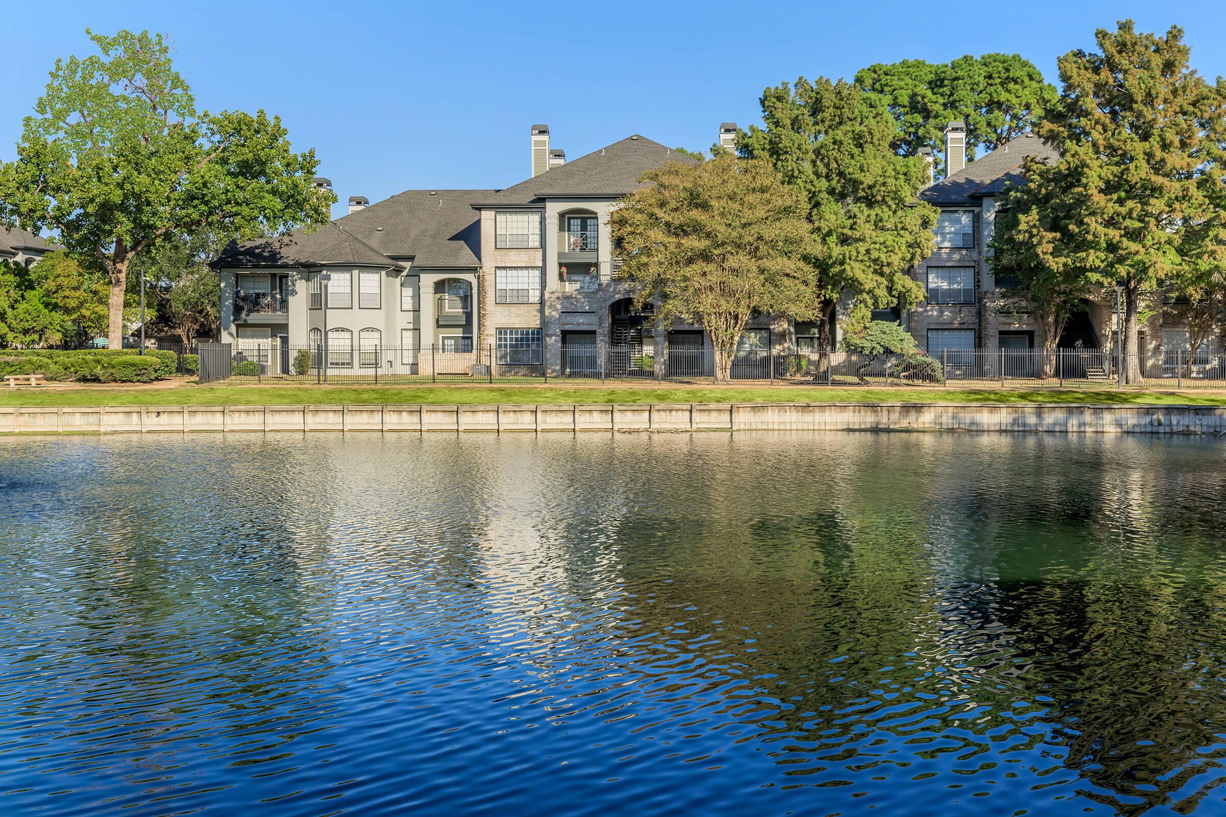 A serene view of a calm lake reflecting the surrounding trees and apartment buildings on a clear day. The buildings feature modern architecture with large windows and balconies, set against a backdrop of greenery, showcasing a peaceful residential landscape.