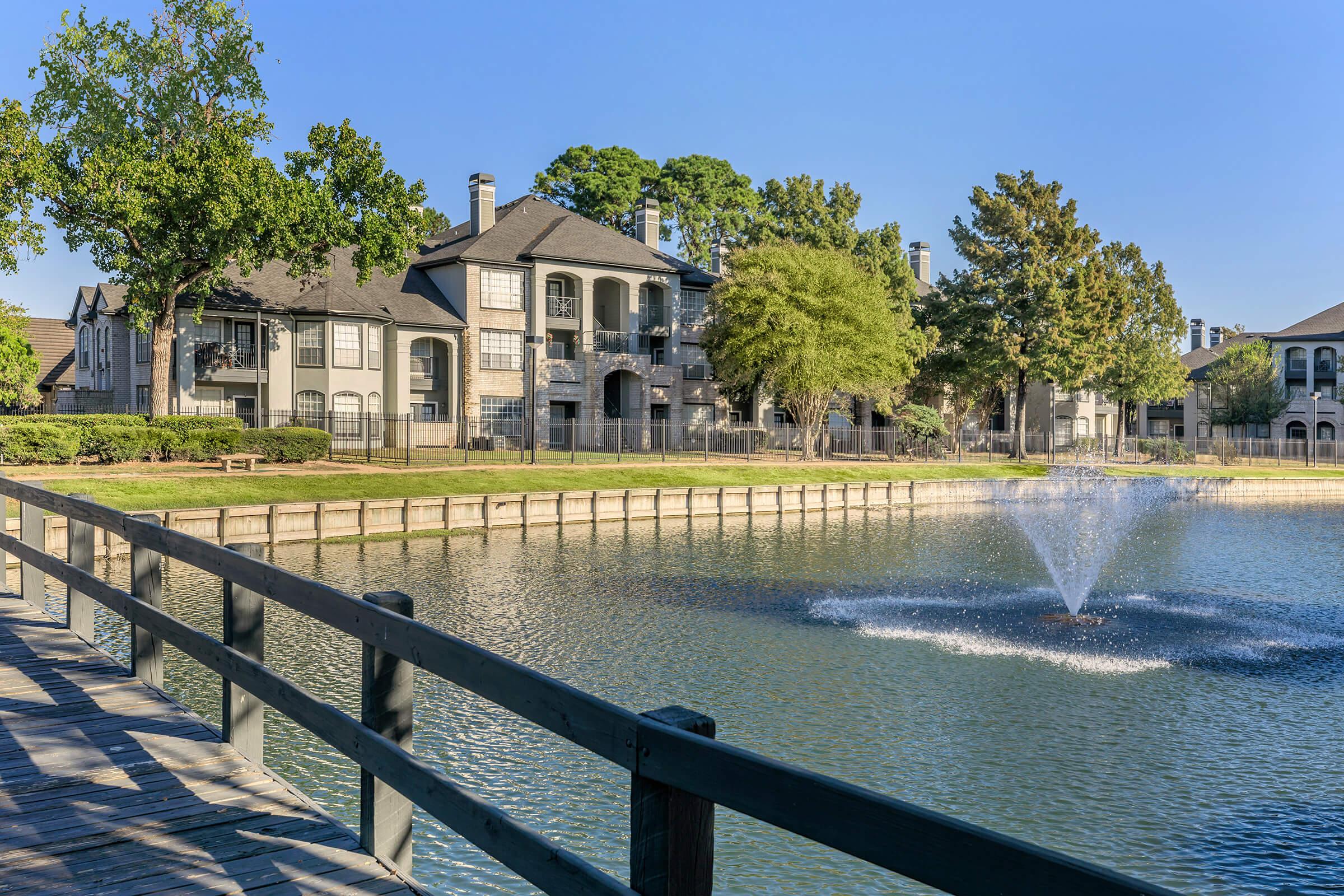 A serene view of a pond with a fountain, surrounded by landscaped greenery and residential buildings. A wooden walkway lines the pond, inviting leisurely strolls. Bright blue skies enhance the peaceful atmosphere of this picturesque setting.