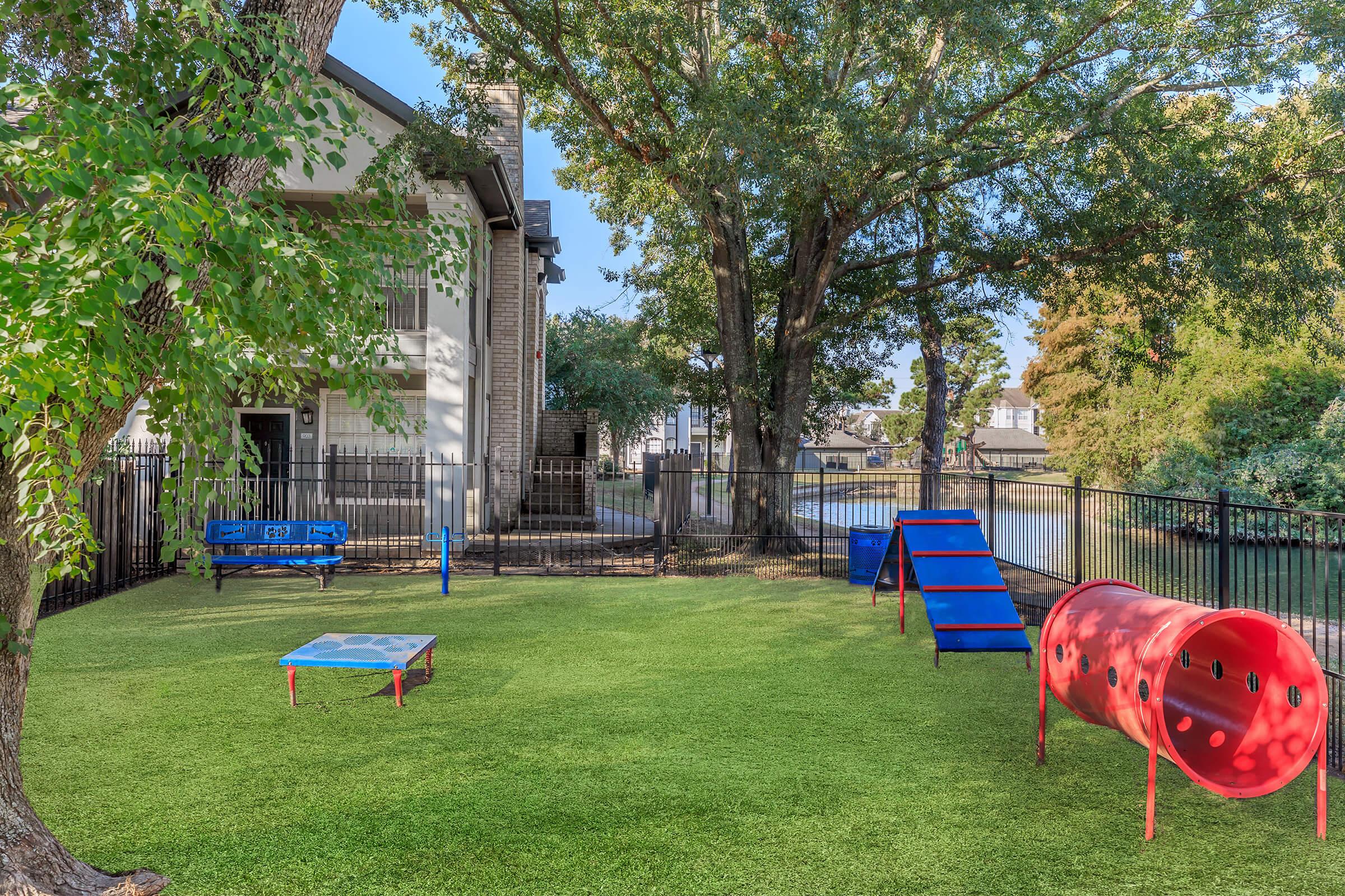 A grassy outdoor play area featuring a blue climbing structure, a red tunnel, and a small blue table. Surrounded by a black fence, trees provide shade. A water view is visible in the background, enhancing the serene atmosphere of this recreational space.