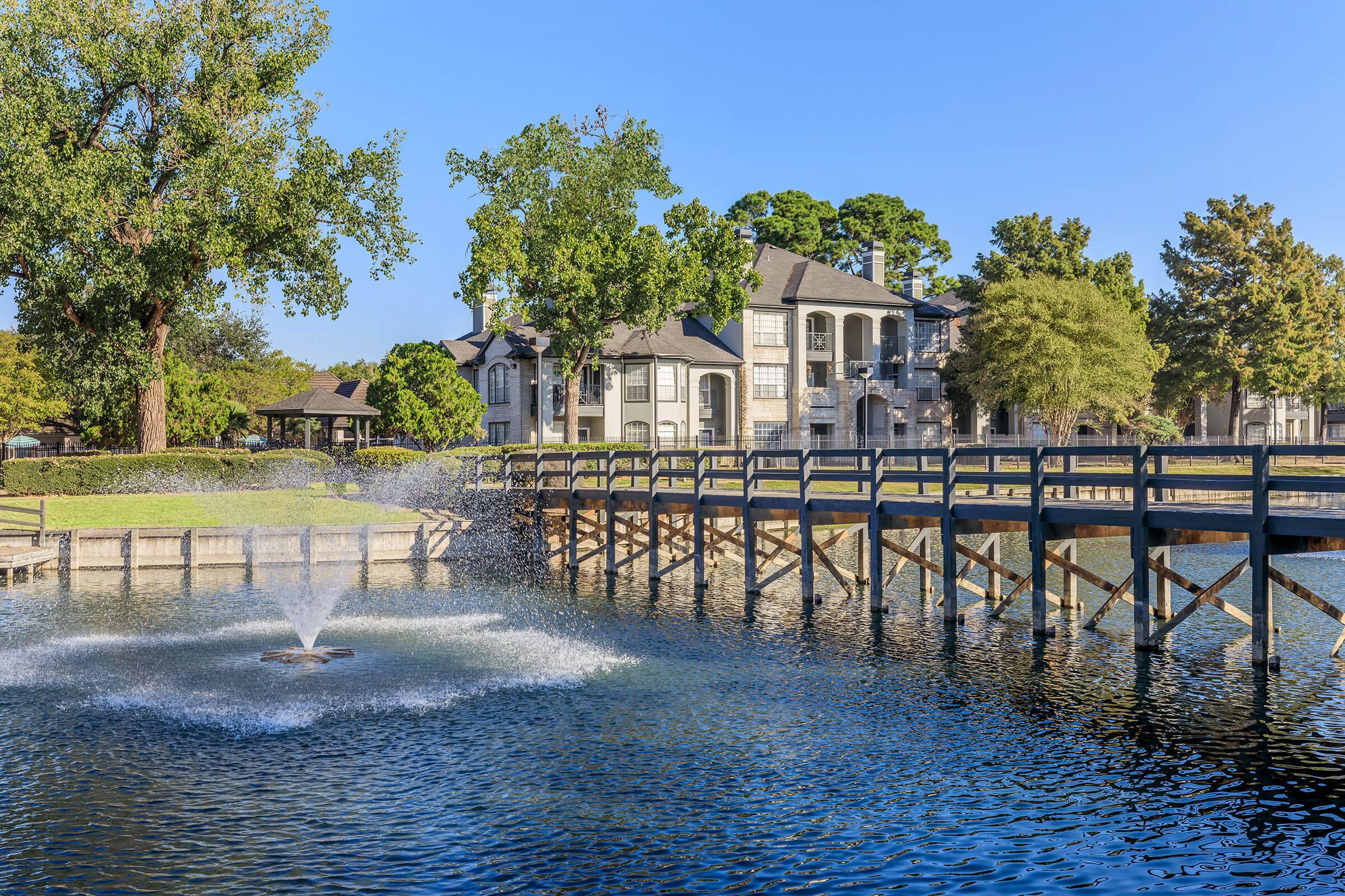 A serene lakeside view featuring a wooden bridge leading to a fountain in the water. Plush greenery surrounds the area, with modern buildings in the background under a clear blue sky, creating a tranquil and picturesque scene.