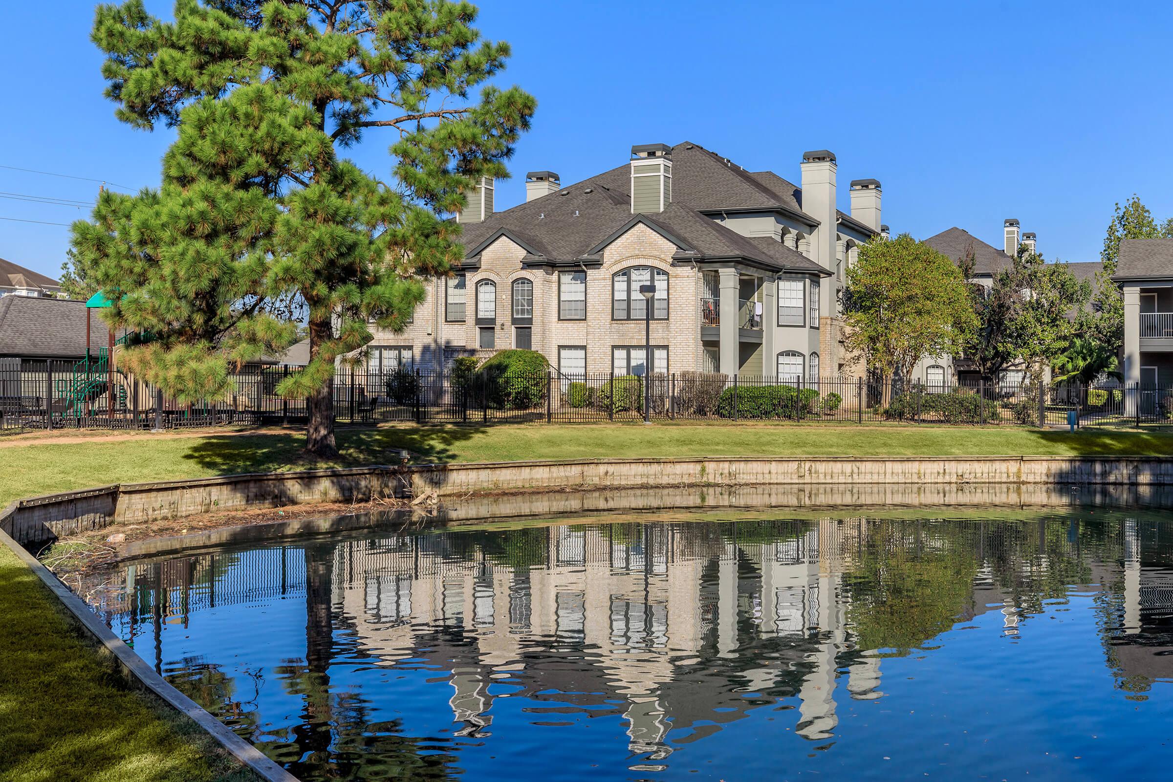 A peaceful landscape featuring a calm pond reflecting a multi-story brick apartment building. Lush greenery surrounds the water, with tall trees and well-maintained grass nearby. The clear blue sky enhances the serene atmosphere of the scene.