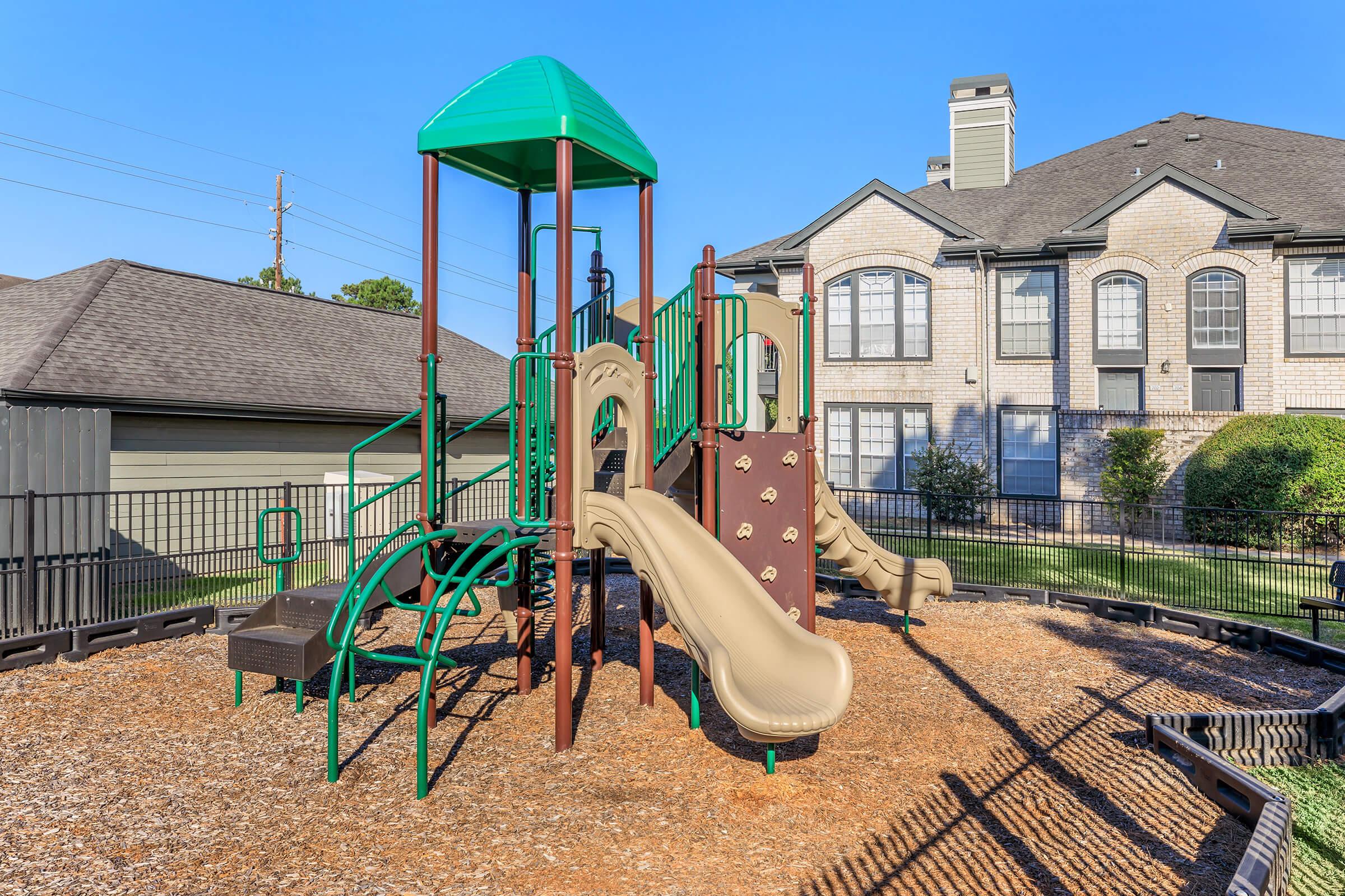 Colorful playground structure with slides and climbing features, situated on a bed of wood chips, surrounded by green grass and a fenced area. In the background, a residential building with large windows and a gabled roof is visible under a clear blue sky.