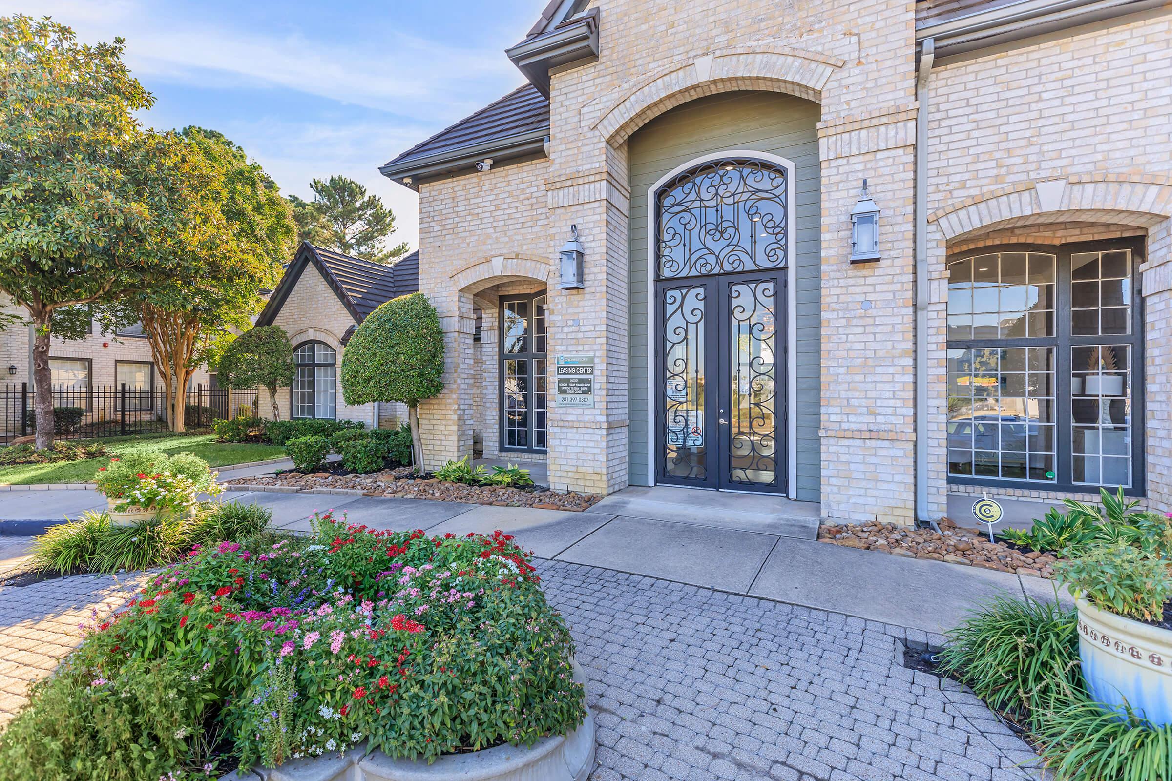 A welcoming entrance to a charming brick building featuring large decorative doors, surrounded by well-maintained landscaping with colorful flowerbeds and trimmed shrubs. The scene captures a bright day with clear skies, highlighting the beauty of the architecture and greenery.
