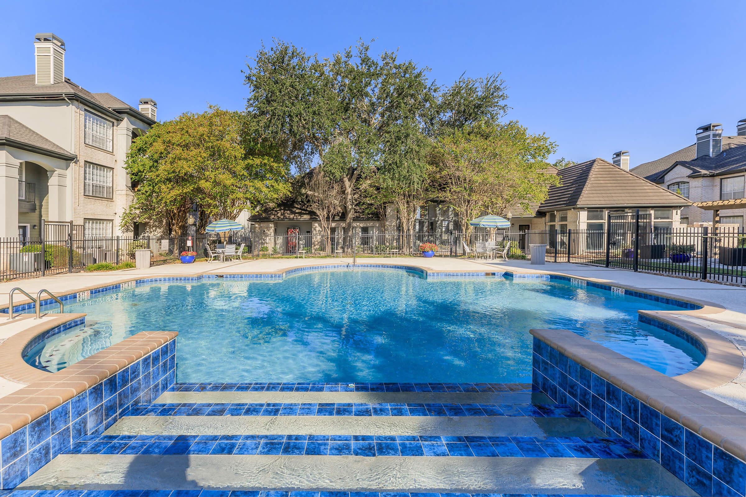 A serene swimming pool surrounded by lush trees and a well-maintained landscape. The pool features a graduated entry with steps, lounge chairs nearby, and a clear blue sky overhead. This inviting outdoor space is perfect for relaxation and leisure.