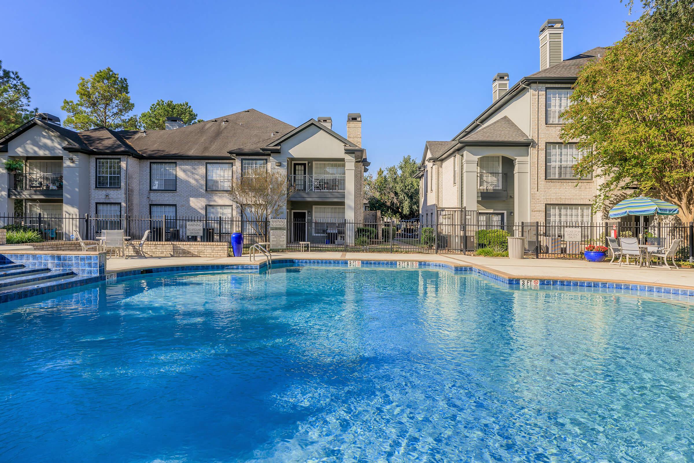 A clear blue swimming pool surrounded by modern apartment buildings. The pool area features lounge chairs, umbrellas, and lush greenery. The scene is bathed in bright sunlight, creating a relaxing and inviting atmosphere.