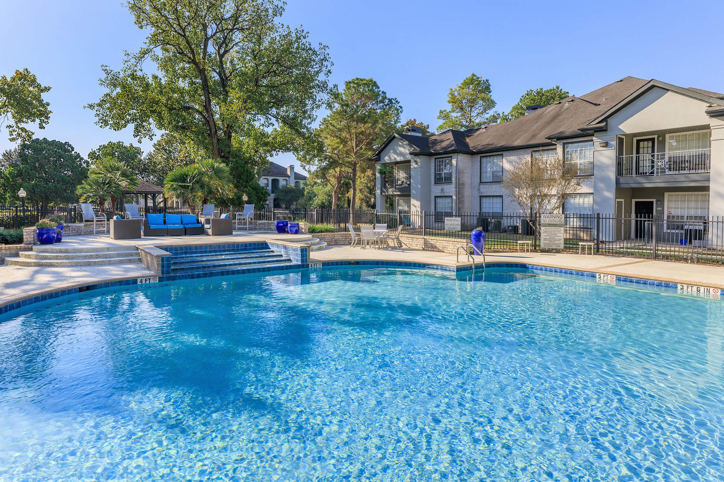 A serene swimming pool surrounded by lush trees and well-maintained greenery. The pool area features steps leading into the water, lounge chairs, and a shaded seating area. Adjacent buildings are visible, creating a relaxing environment perfect for leisure and enjoyment. Bright blue sky completes the picturesque scene.