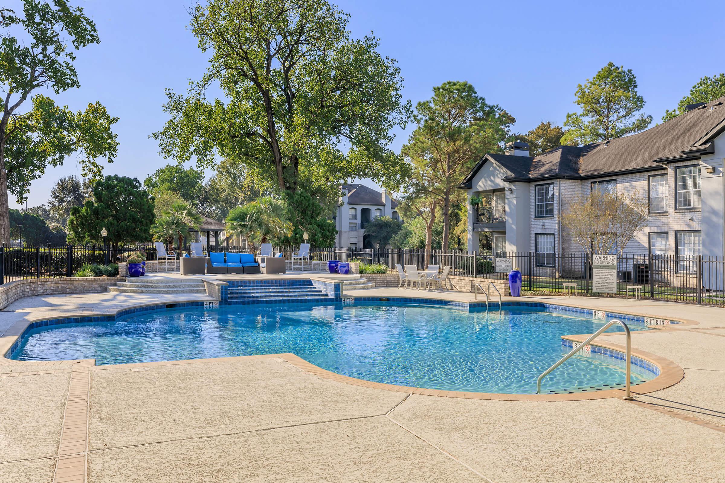 A sparkling outdoor swimming pool surrounded by a concrete deck, nestled among trees and greenery. Nearby, lounge chairs and a seating area are visible, along with an apartment building in the background under a clear blue sky.