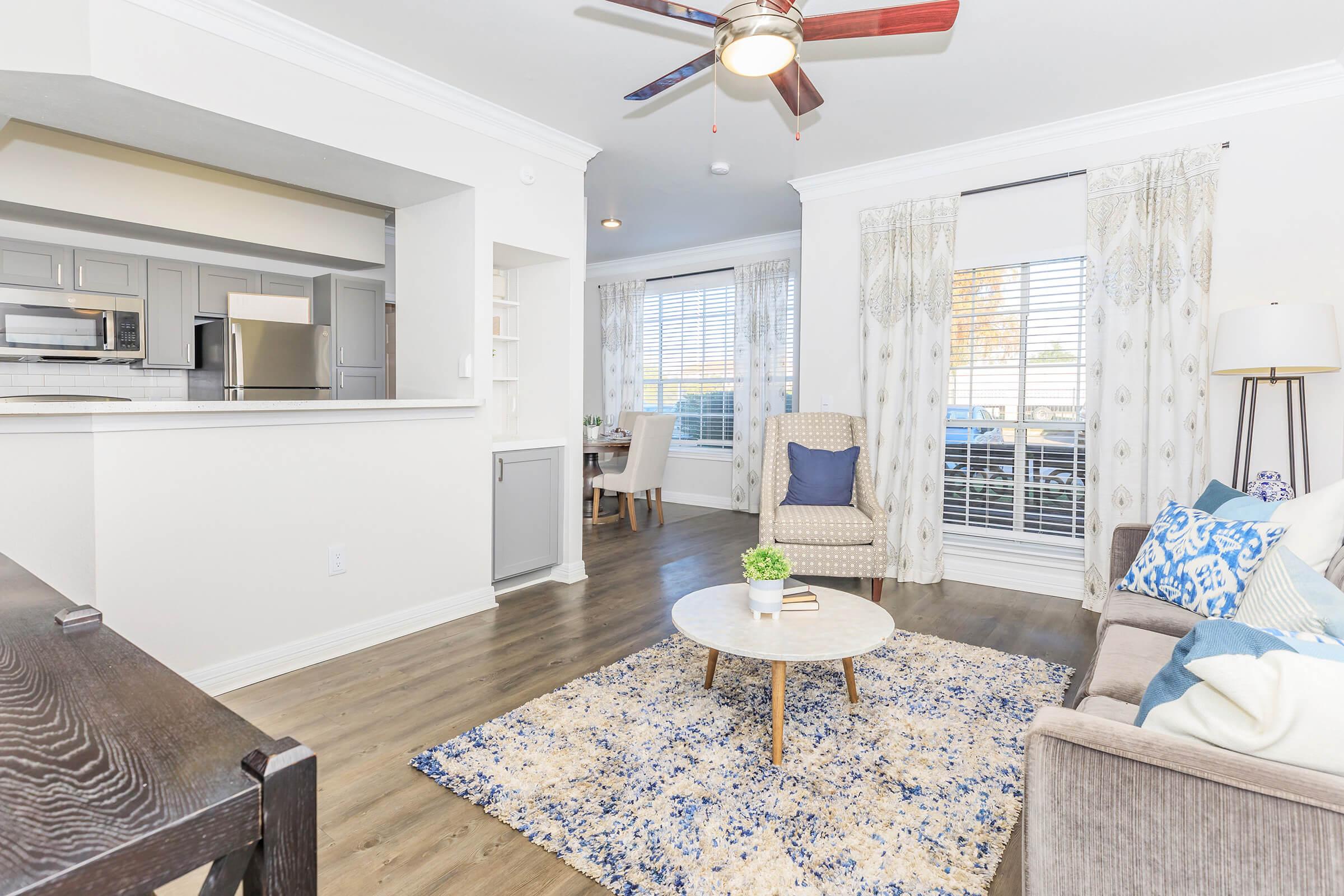 A modern living room featuring a cozy beige sofa adorned with blue and white pillows. There’s a round coffee table in the center on a patterned area rug, and a ceiling fan overhead. Light streams in through large windows with sheer curtains, complementing the neutral color scheme and inviting atmosphere.