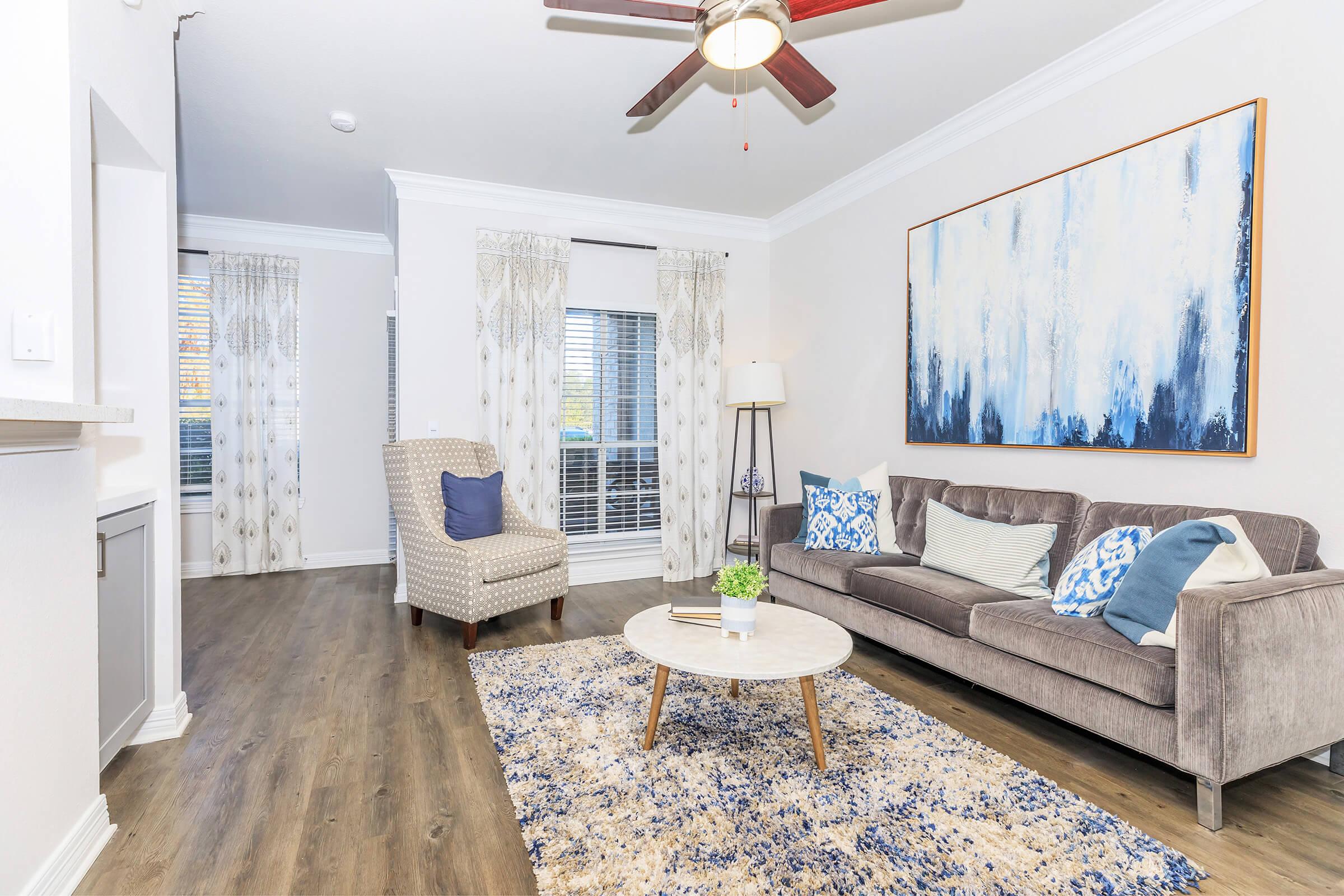 A modern living room featuring a gray sofa with blue and white decorative pillows, a round coffee table with a marble top, and a patterned rug. A cozy armchair and a floor lamp are positioned near large windows with curtains. The walls are painted light, and there is a contemporary artwork above the sofa.