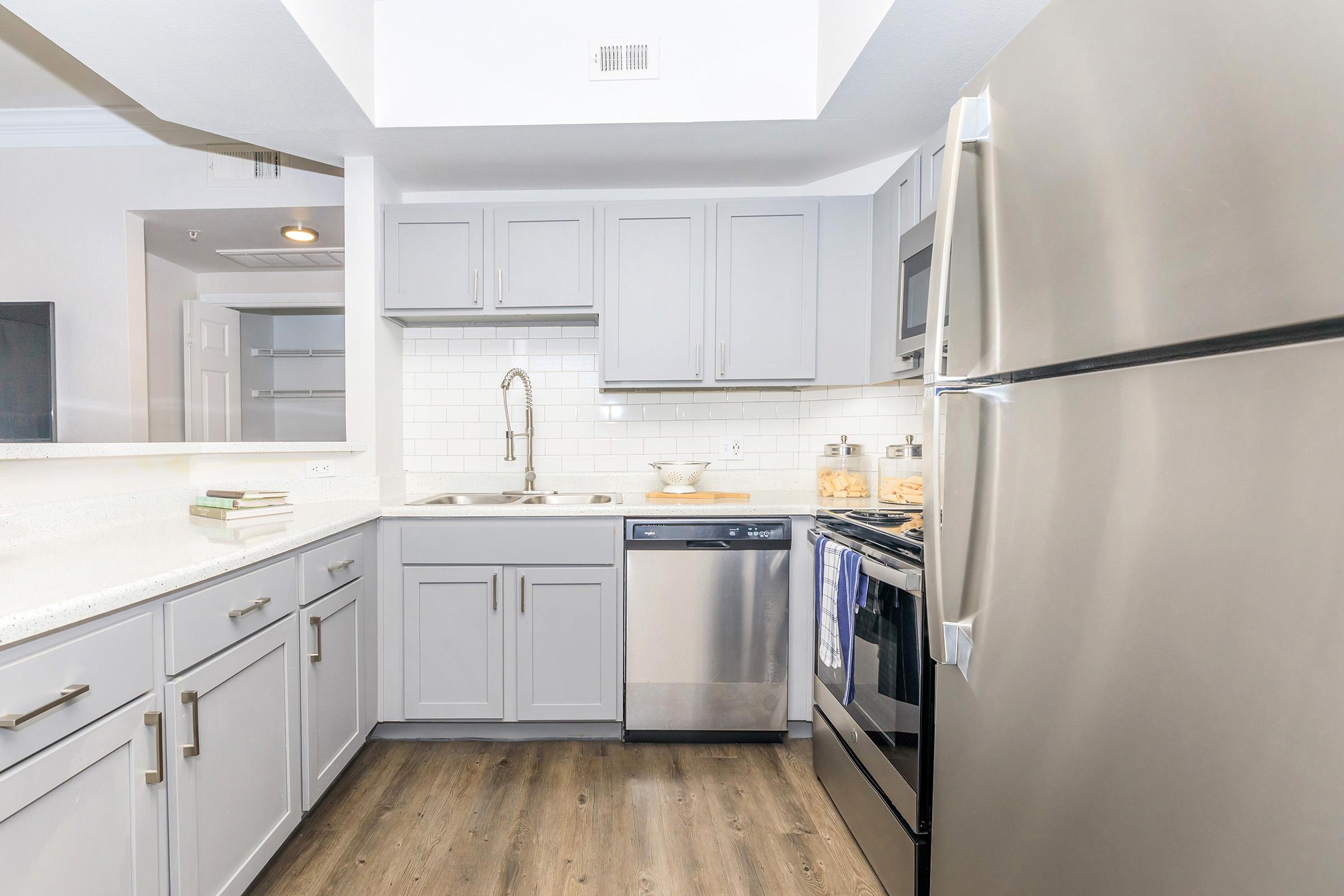 A modern kitchen featuring light gray cabinetry, a stainless steel refrigerator, and a dishwasher. The countertop is a light-colored material, and there is a stainless steel sink with a pull-down faucet. A window provides natural light, and the flooring is a wood-look design, creating a bright and contemporary atmosphere.
