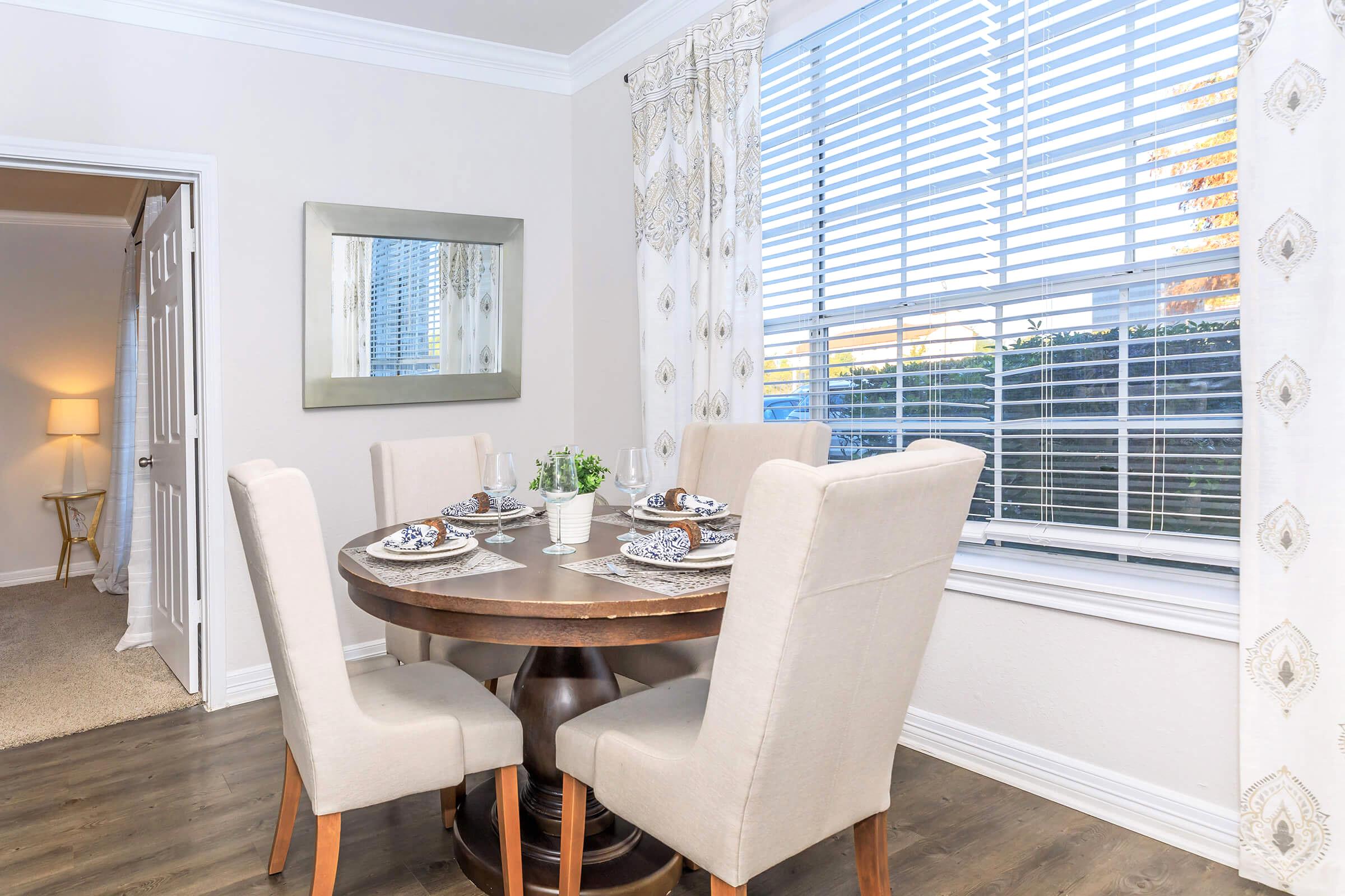 A cozy dining area featuring a round wooden table set for four with plates and cutlery. The space has light-colored walls, a mirror on the wall, and sheer window treatments allowing natural light to fill the room. A decorative plant sits in the center of the table, creating an inviting atmosphere.