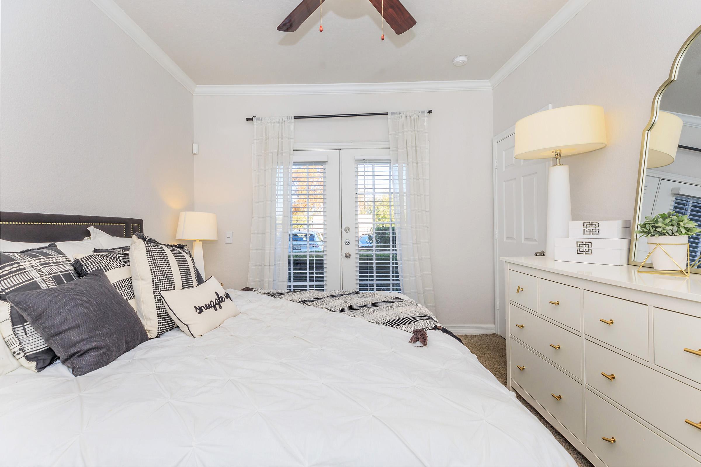A cozy bedroom featuring a neatly made bed with black and white patterned pillows and a white comforter. There’s a stylish dresser with golden handles, a decorative lamp, and a small potted plant on top. Natural light streams in through the open double doors, complemented by sheer white curtains.