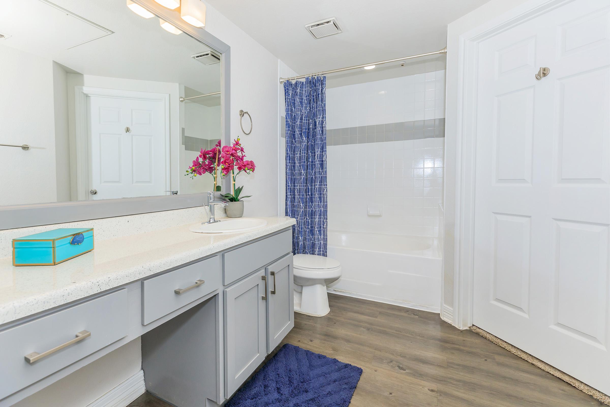 A well-lit bathroom featuring a white bathtub with a blue shower curtain, a modern vanity with a granite countertop, and a mirror. A small blue box sits on the countertop, and a purple rug adds color to the wood-like flooring. Fresh flowers in a vase enhance the decor.