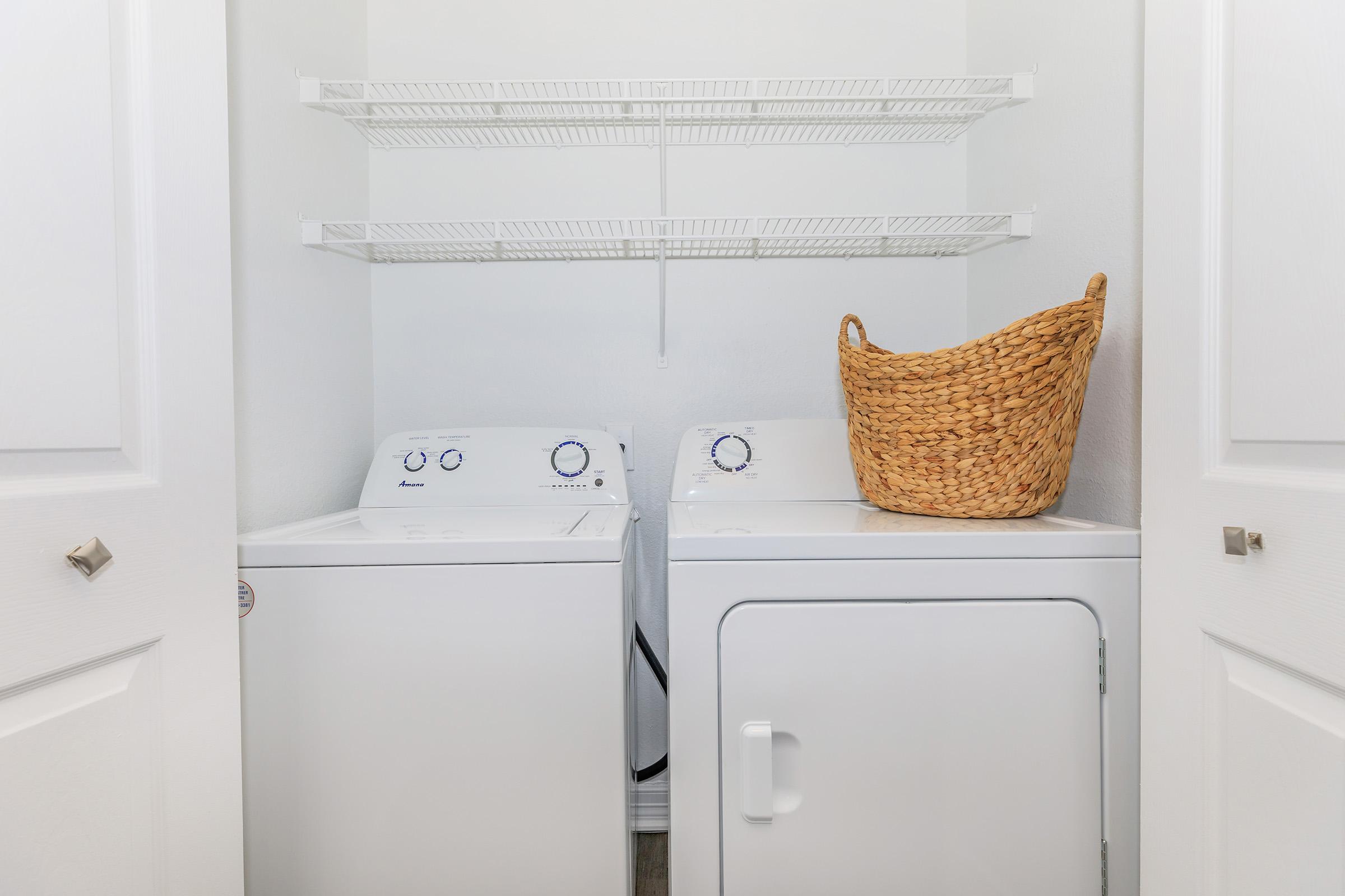 A clean laundry area featuring a white washing machine and dryer side by side, with a simple wire shelving unit above and a woven basket beside them. The space is bright and tidy, with white walls and closed doors in the foreground.
