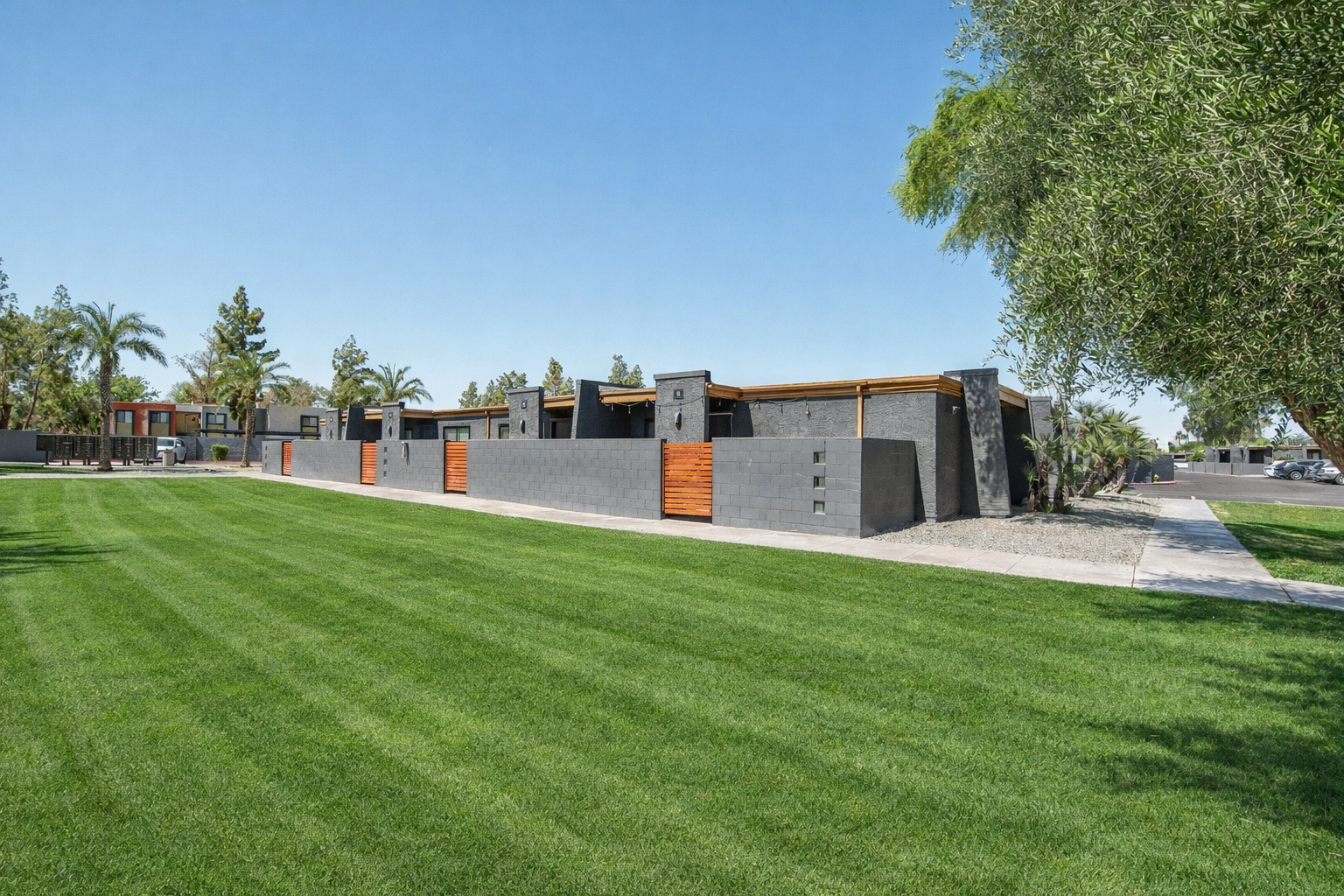 A modern, single-story building with a mix of gray and wooden elements surrounded by lush green grass. Palm trees and other landscaping adorn the area, with a clear blue sky overhead. The design features clean lines and a contemporary aesthetic, creating an inviting outdoor space.