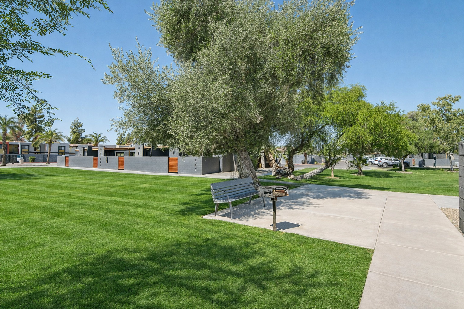 A well-maintained park area featuring lush green grass, a concrete walkway, a bench, and a barbecue grill. Surrounding the park are trees and modern buildings, with parking spaces visible in the background under clear blue skies.