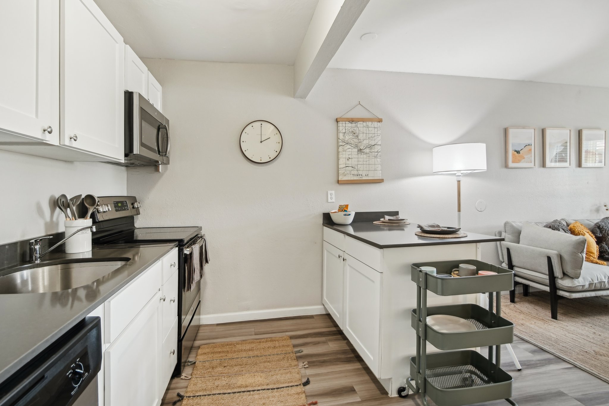 A modern kitchen and living area featuring white cabinets, a stainless steel stove and microwave, a wall clock, and a decorative wall hanging. A cart with dinnerware is in the foreground, while a cozy gray sofa is arranged nearby, creating a stylish and functional space.