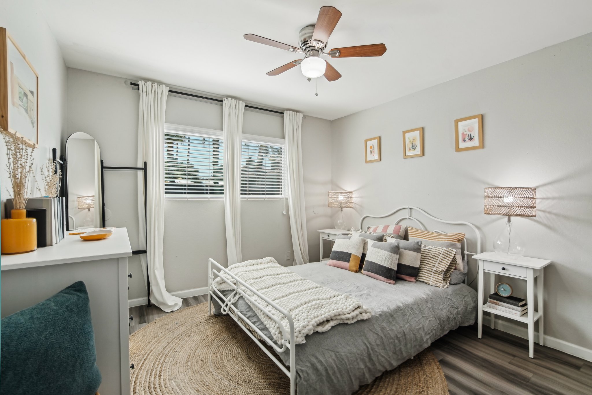 A cozy bedroom featuring a white metal bed with colorful throw pillows, a textured blanket, and a round rug. There's a modern nightstand with a lamp, a dresser, and large windows with sheer curtains allowing natural light. Wall art adds a personal touch to the inviting space.