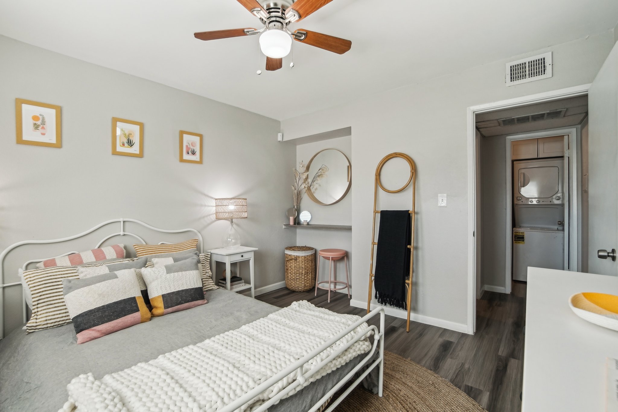 A cozy bedroom featuring a white metal bed with decorative pillows, a lamp, and framed art on the walls. There is a round mirror, a wooden rack, and a small table with two stools. The room has neutral colors, wooden flooring, and a laundry area visible in the background.