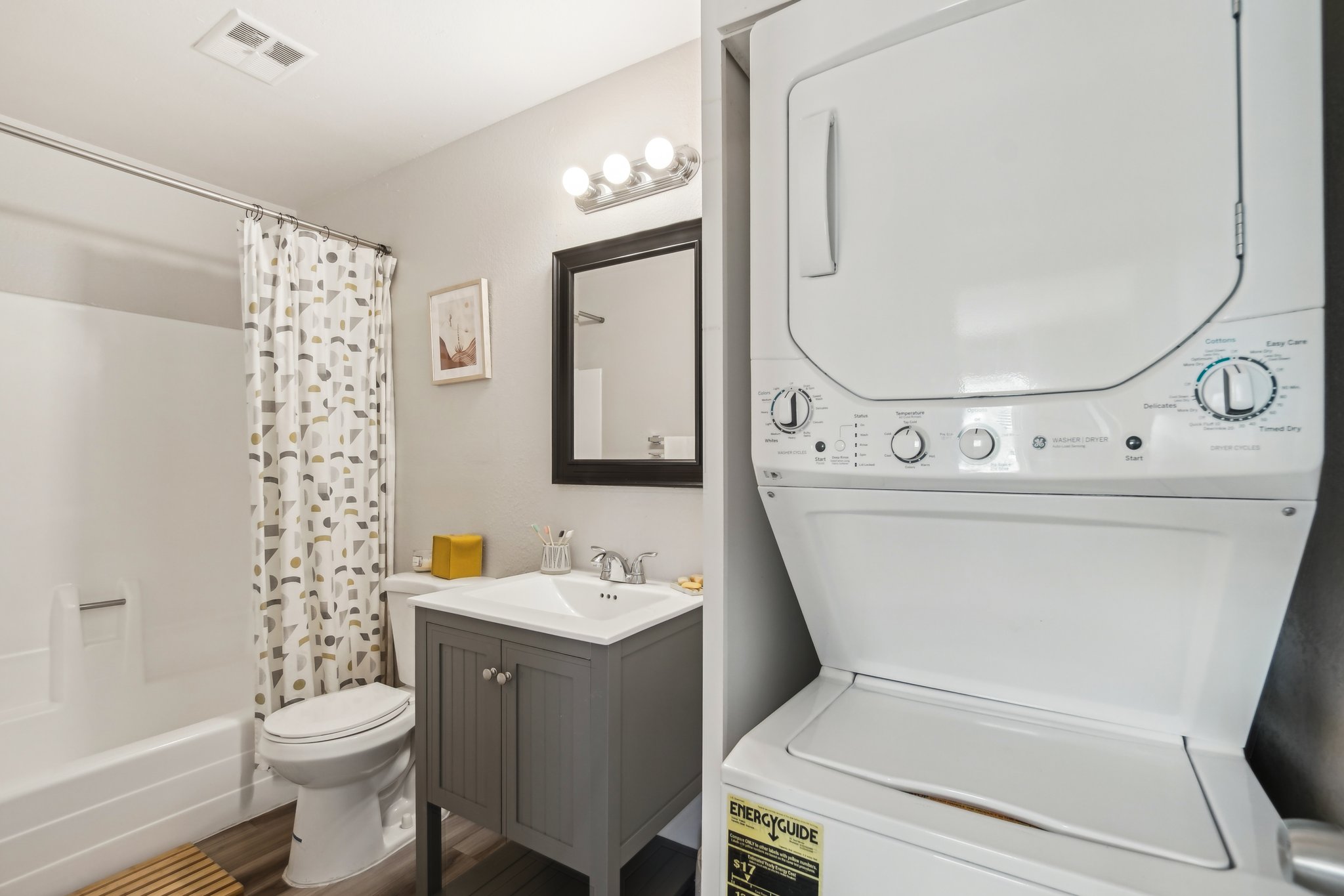 A modern bathroom featuring a shower with a curtain, a toilet, a sleek gray vanity with a sink, and a mirror. On the side, there is a stacked washer and dryer unit. The decor includes a framed picture and a yellow accent piece, with wooden flooring and soft lighting.