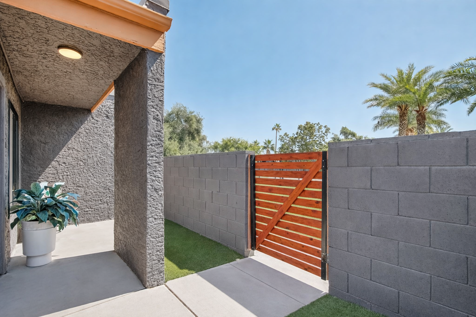 A view of a modern outdoor space featuring a concrete wall with a wooden gate, lush green grass, and a potted plant. The bright blue sky and palm trees in the background create a tropical atmosphere, inviting relaxation. The area is well-lit, showcasing the contemporary design.