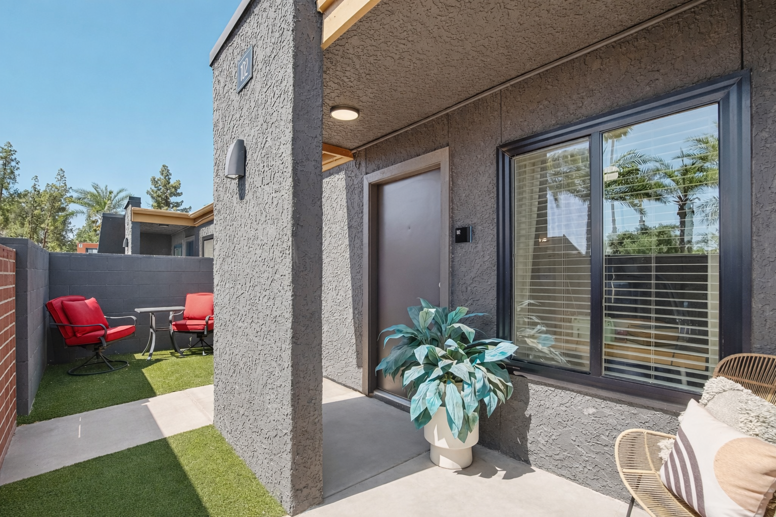A modern exterior view of a home featuring a small patio area with red chairs and a table. There is a large potted plant near the entrance, a door with a number, and windows with blinds. The surrounding area includes green grass and palm trees in the background under a clear blue sky.