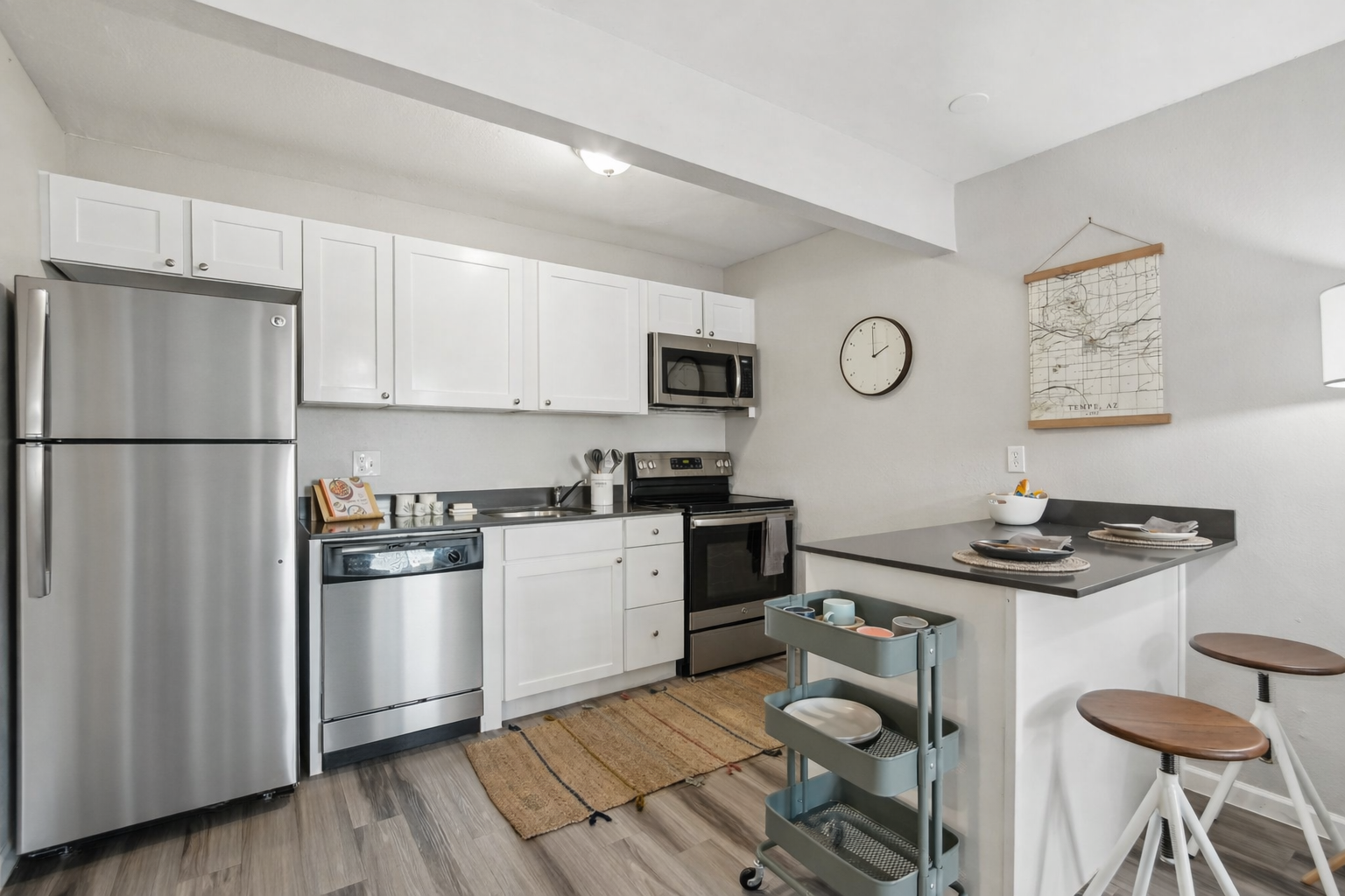 Modern kitchen featuring stainless steel appliances, including a refrigerator, microwave, and oven. White cabinets and countertops complement a minimalist design. There's a kitchen island with bar stools and a rolling cart with dishware. A wall clock and a framed map add decorative touches.