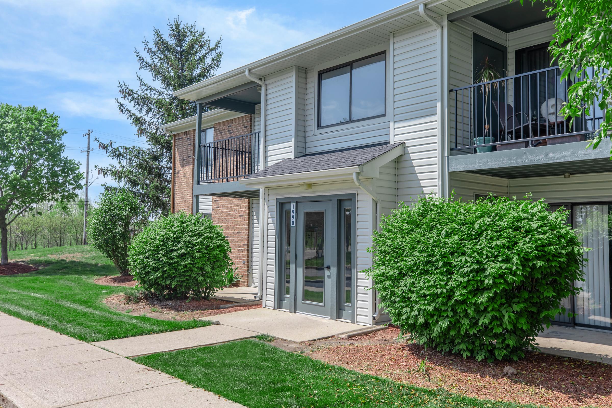 Exterior view of a two-story apartment building with a mix of brick and siding. The entrance features a glass door and is flanked by two large green bushes. Sidewalk leading up to the door, with well-maintained landscaping and trees in the background under a clear blue sky.