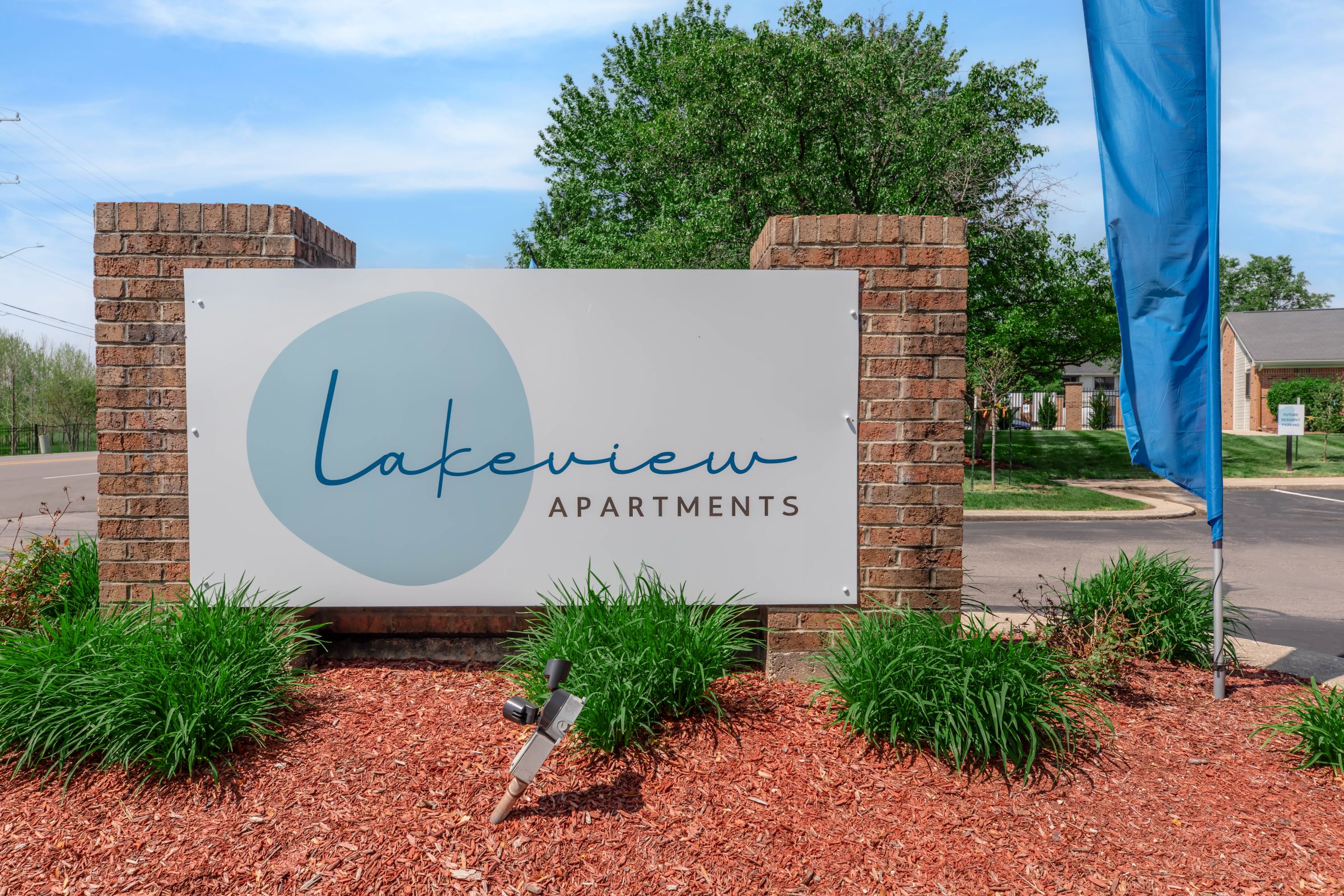 Sign for Lakeview Apartments displayed on a brick structure, featuring a blue circular logo and modern typography. The surrounding area includes green landscaping, with small shrubs and mulch, along with a banner pole on the left. Visible road and trees in the background.