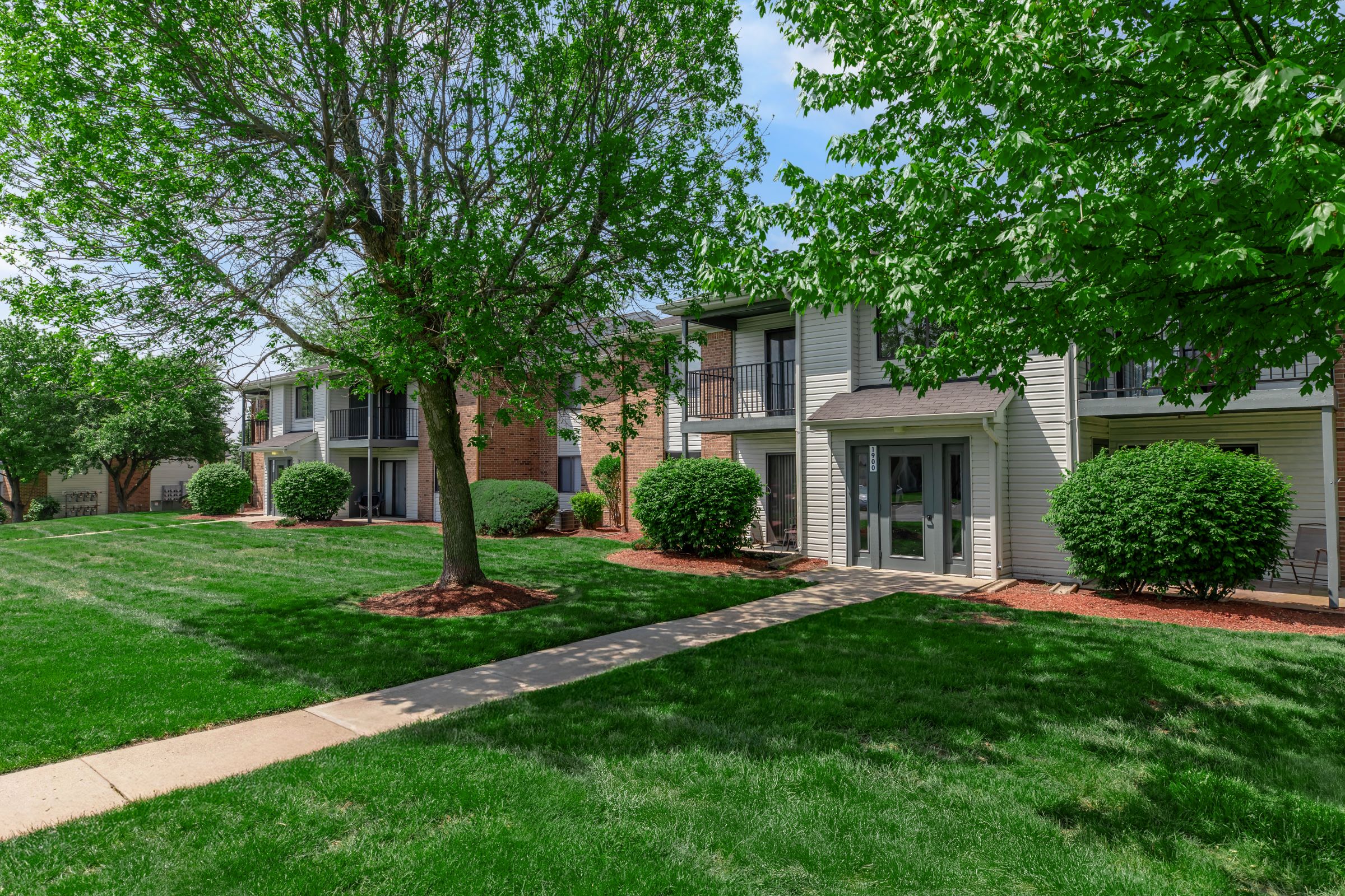 A landscaped courtyard featuring several apartment buildings, surrounded by lush green grass and small shrubs. There are pathways leading through the open space, with a few trees providing shade against a clear blue sky. The setting is peaceful and well-maintained, ideal for outdoor activities.