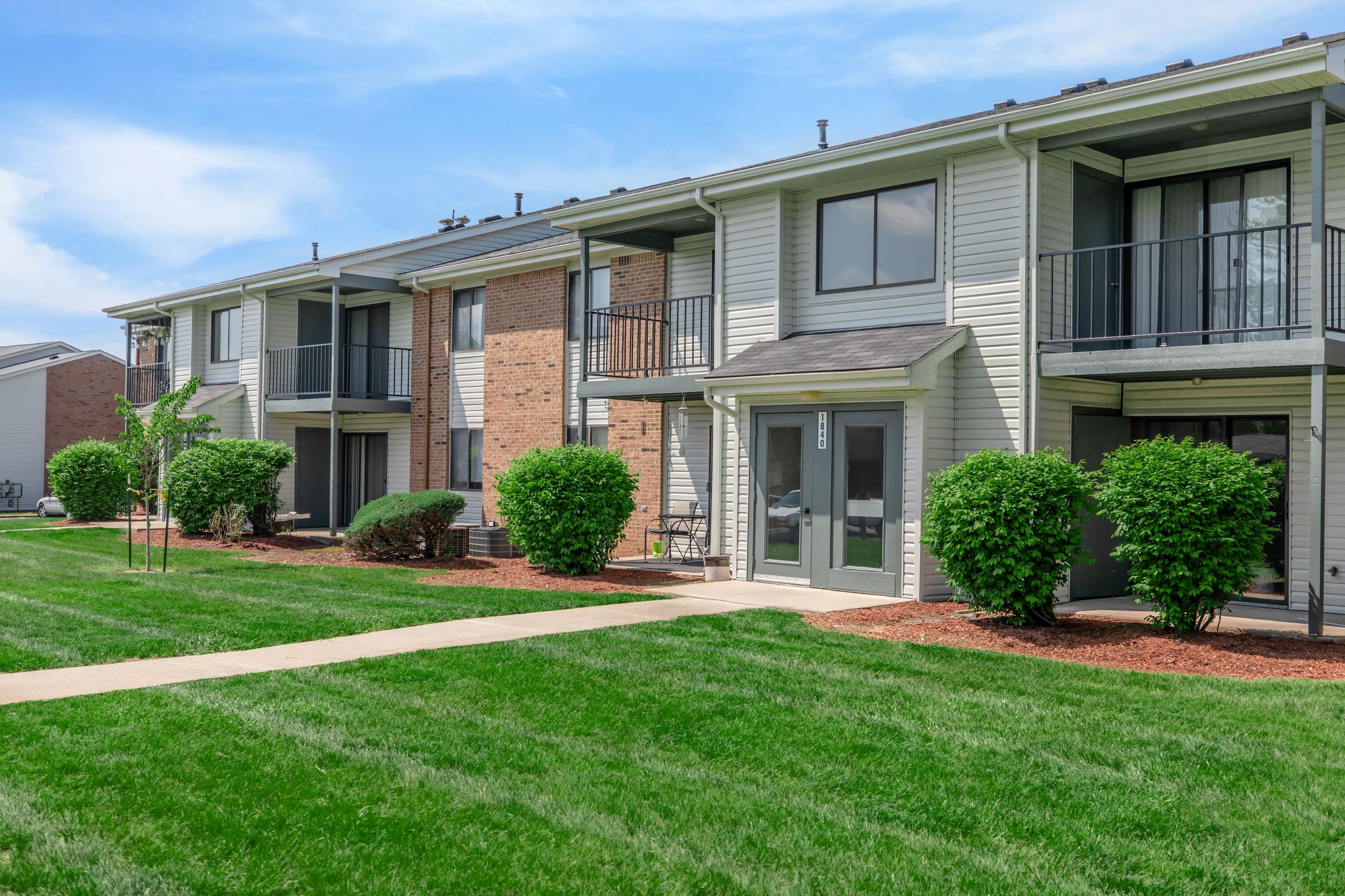 A well-maintained apartment complex featuring two-story buildings with a mix of brick and light-colored siding. Each unit has a small patio or balcony and is surrounded by neatly landscaped lawns and shrubs. The walkway leads through the grassy area, creating an inviting atmosphere.