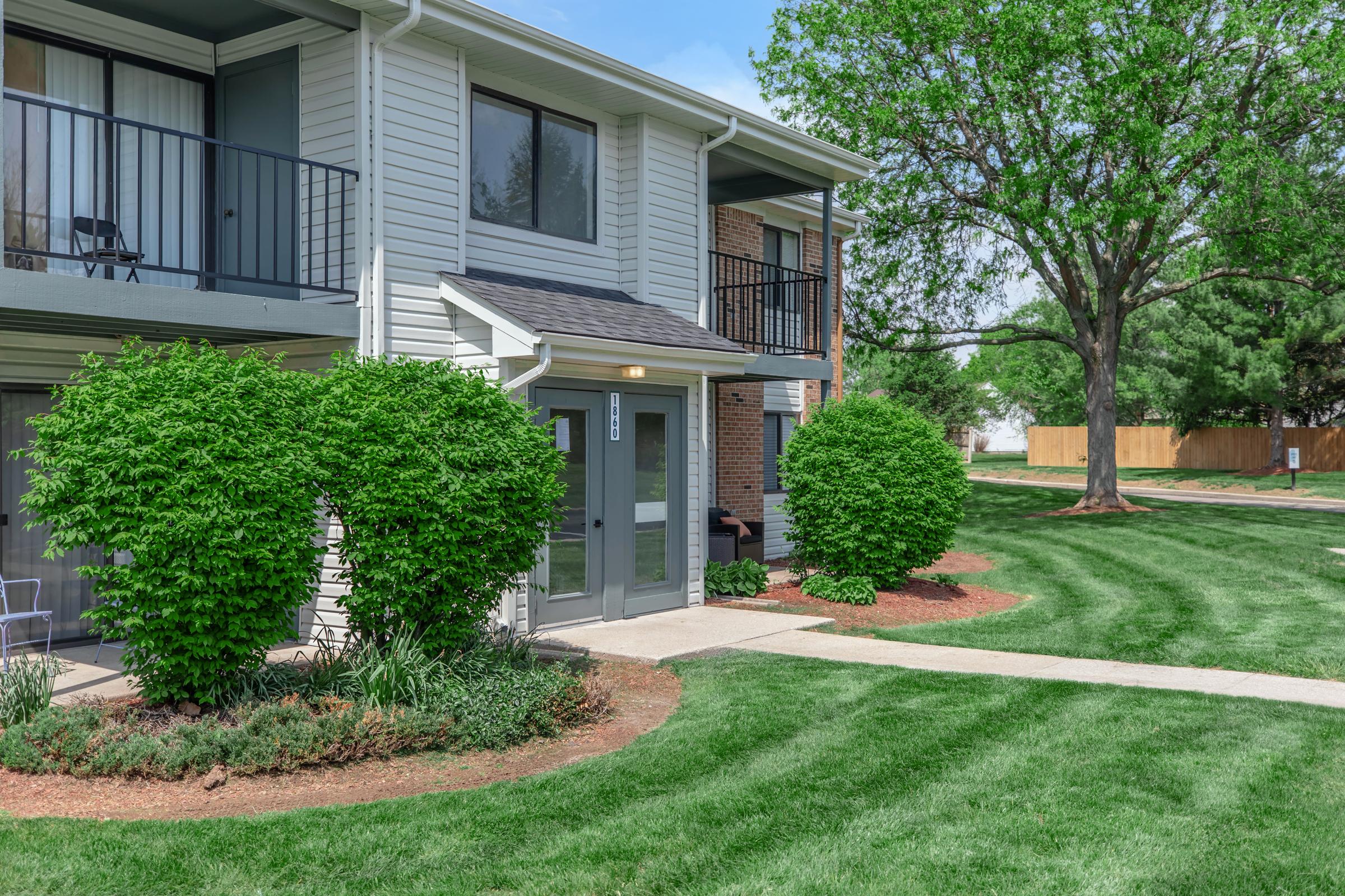 A well-maintained apartment complex features two buildings with light-colored siding. The entrance is highlighted by a small patio, surrounded by neatly trimmed shrubs and flower beds. Lush green grass and trees create a welcoming outdoor space, with a pathway leading up to the doors of the units.