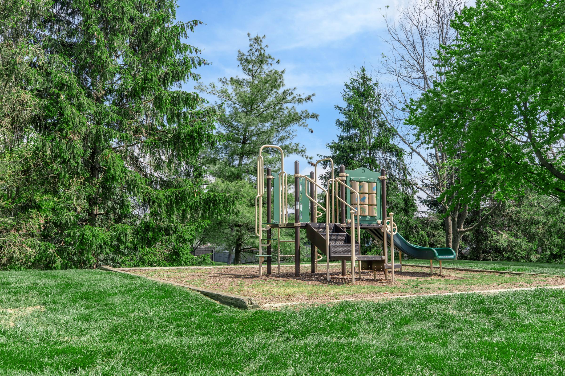 Playground equipment in a park, featuring a slide, climbing structure, and steps, surrounded by green grass and trees under a blue sky. The area is designed for children to play and explore.