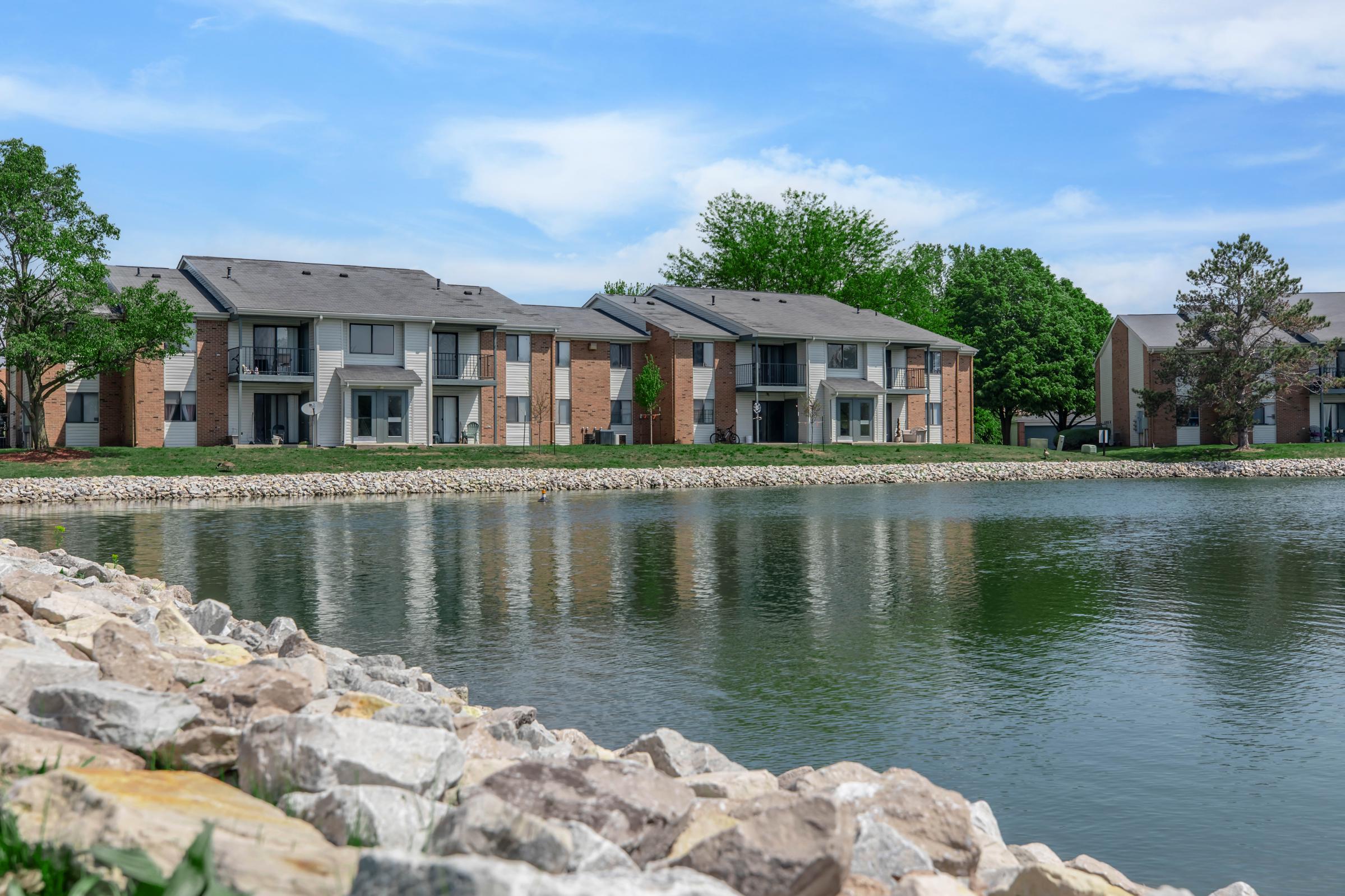 A serene view of apartment buildings lined along a calm lake. The foreground features a rocky shoreline, while lush greenery and trees provide a natural backdrop under a blue sky with scattered clouds. The scene conveys a peaceful residential atmosphere.