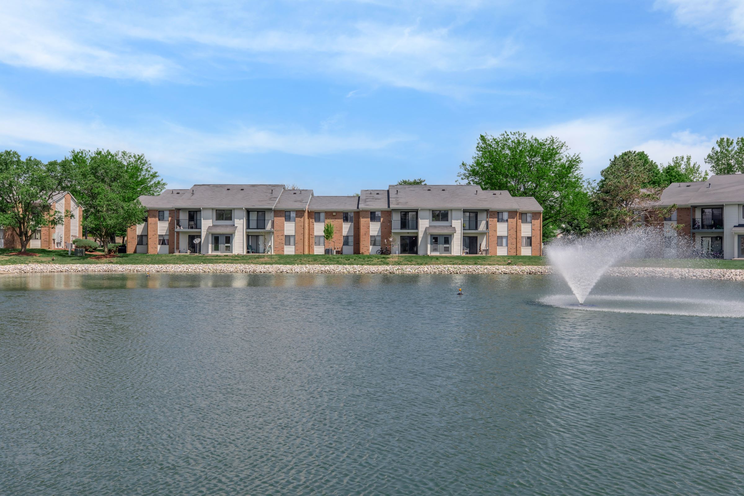 A serene view of a pond with a fountain, surrounded by lush greenery and multi-story apartment buildings in the background. The sky is clear with some clouds, creating a peaceful and inviting atmosphere.