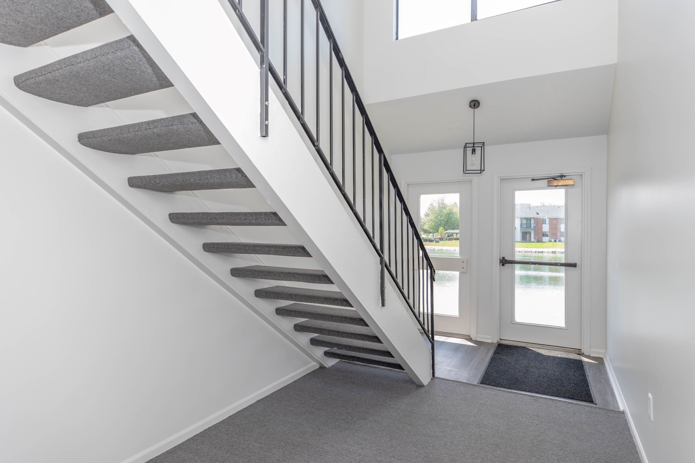 A modern interior entryway featuring a staircase with gray carpeting and a black metal railing. Natural light floods the space through a large window. There's a door leading outside, with a view of a lake and green surroundings. The floor is a mix of carpet and laminate, adding to the contemporary feel.