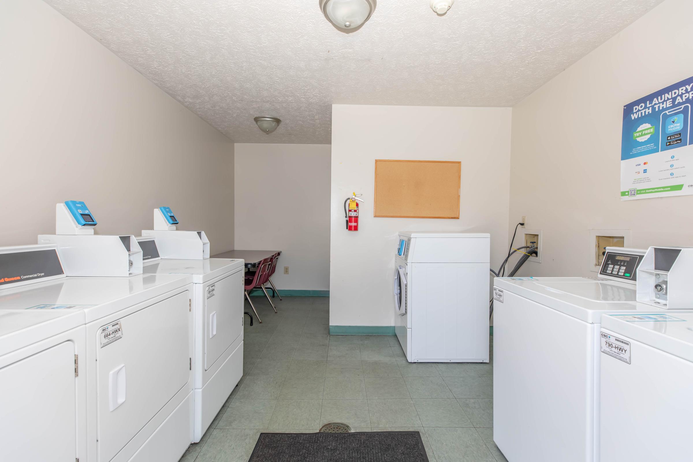 A clean laundry room featuring several white washing machines and dryers lined up against the walls. There is a small table and chairs in the corner, a fire extinguisher on the wall, and a bulletin board. The floor is tiled, and the walls are painted in light colors, creating a bright and functional space.