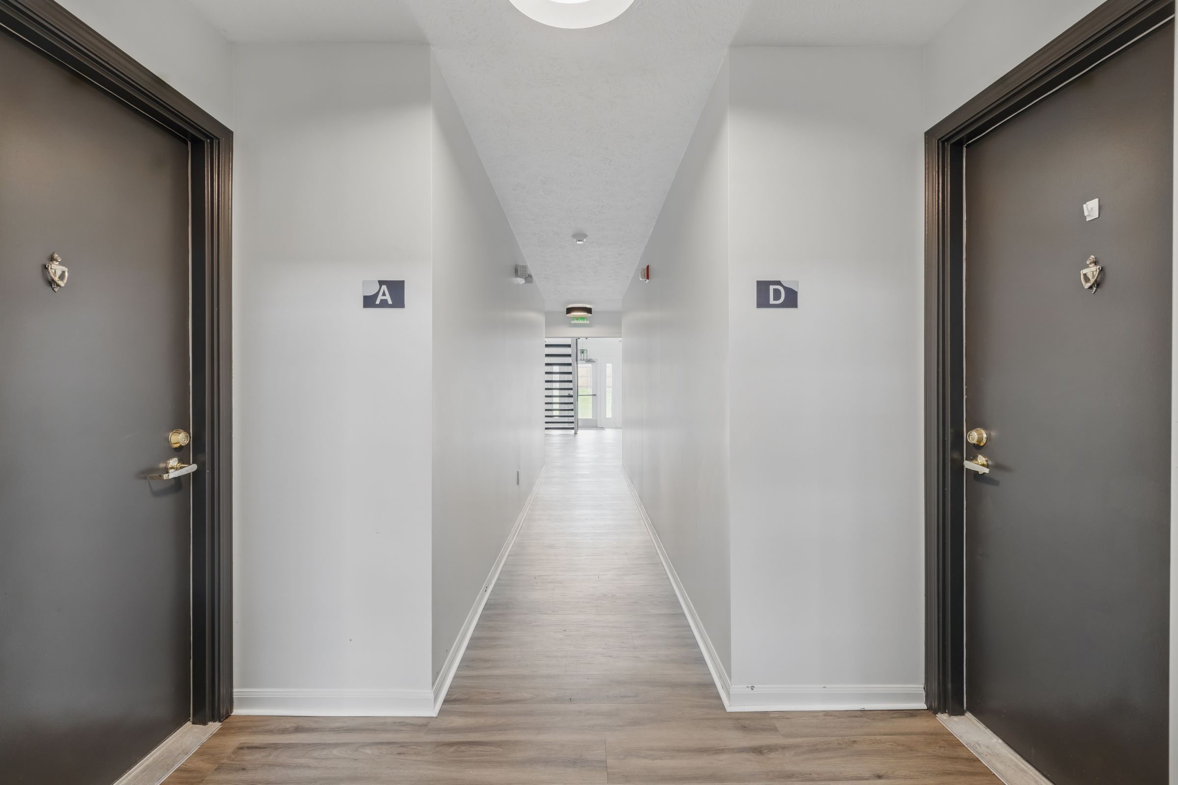 A well-lit corridor with doors marked A and D on either side. The walls are painted white, and the flooring is a warm wood finish. A staircase is visible at the end of the hallway, leading to another level. The space has a modern and clean aesthetic.