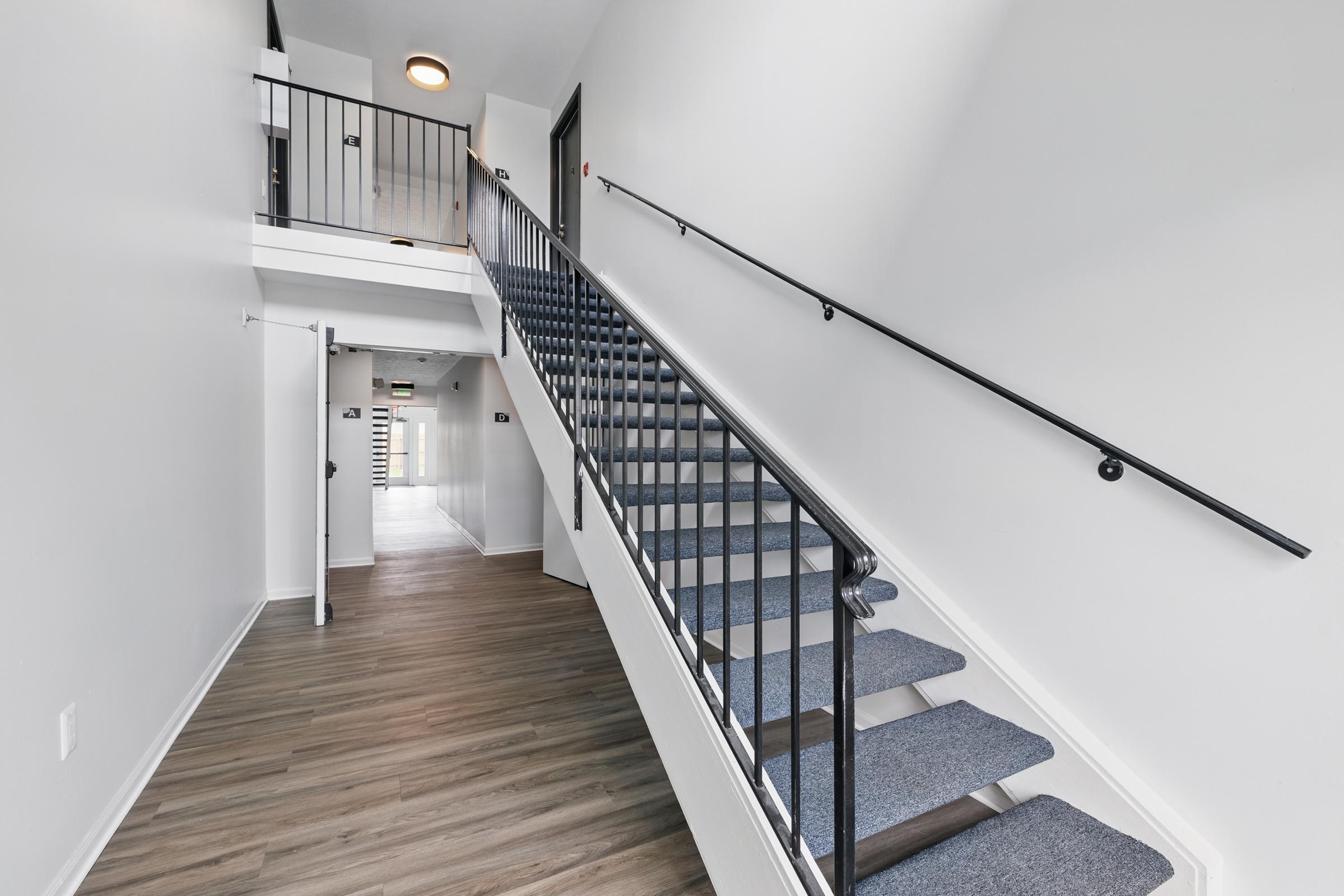 A spacious, well-lit interior staircase with black railings, leading to an upper floor. The walls are painted white, and the floor features light wooden laminate. The staircase is carpeted in blue, with a door visible at the back and additional doors on the upper level. Natural light comes from a ceiling fixture.