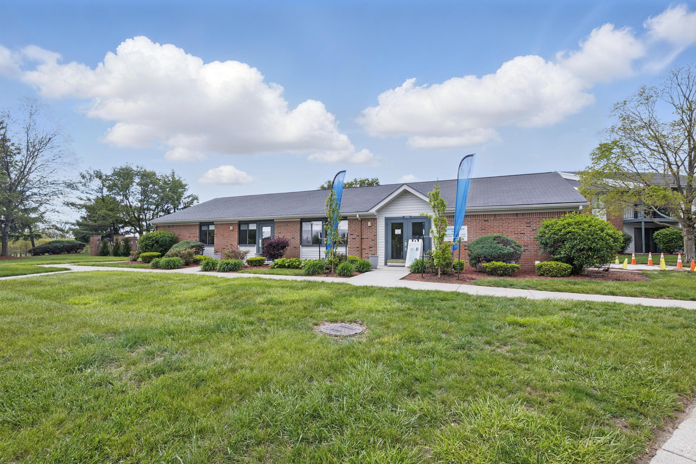 Single-story brick building with a gray roof, surrounded by green grass and shrubs. A path leads to the entrance, which features two blue flags. In the background, trees and more greenery are visible under a bright blue sky with fluffy white clouds.