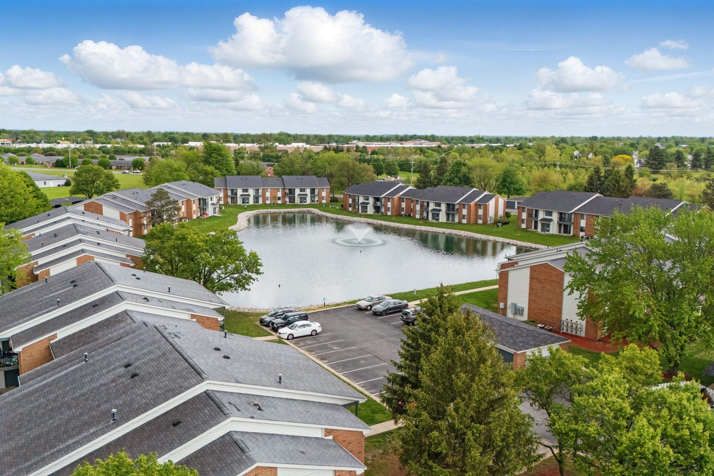 Aerial view of a residential complex surrounding a serene pond with a central fountain. Lush green trees and well-maintained lawns are seen around the buildings, which have a mix of brick and siding exteriors. A parking area is visible, contributing to the peaceful suburban atmosphere.