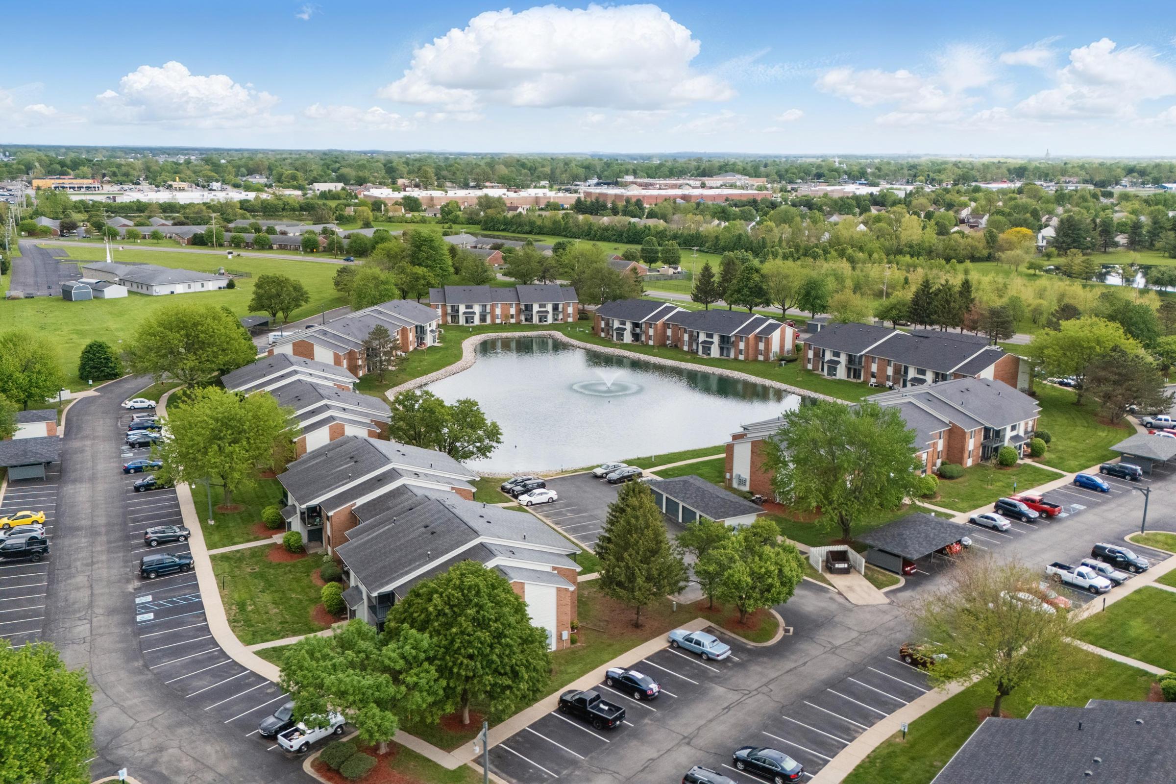 Aerial view of a residential complex featuring multiple buildings arranged around a circular pond. The pond has a fountain in the center, surrounded by green lawns and trees. Parking spaces are visible, with cars parked along the pathways. The background includes additional greenery and suburban structures.