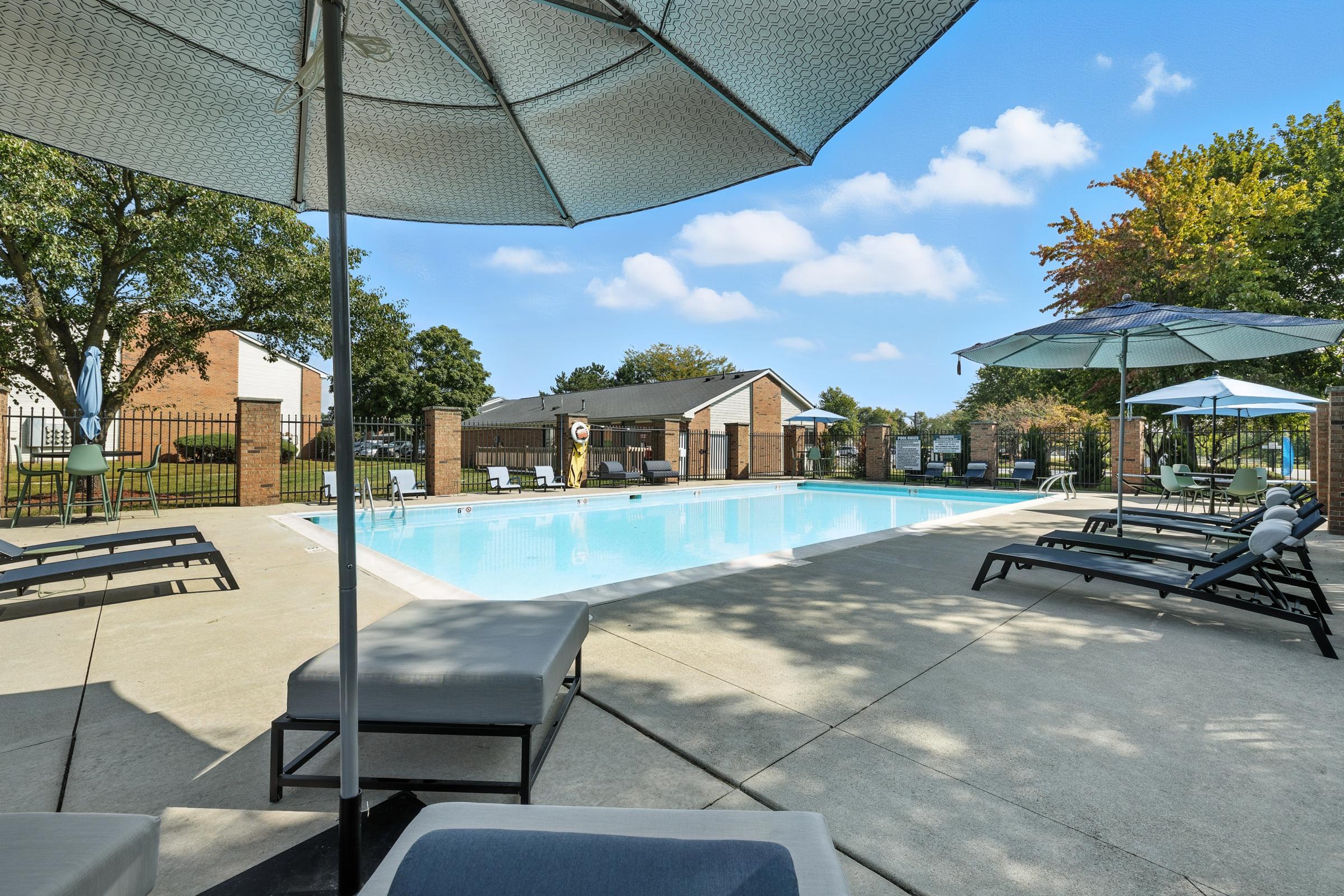 A clear, inviting swimming pool surrounded by lounge chairs and umbrellas. The pool area features a well-maintained concrete deck, with green trees and a few buildings in the background under a blue sky with fluffy clouds.